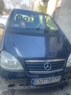 Samochód marki Mercedes-Benz, ciemny kolor, widok z przodu. Widoczny emblemat na atrapie chłodnicy, tablica rejestracyjna KNT 18173. Na przedniej szybie naklejka z napisem 001. W tle budynki i drzewa.