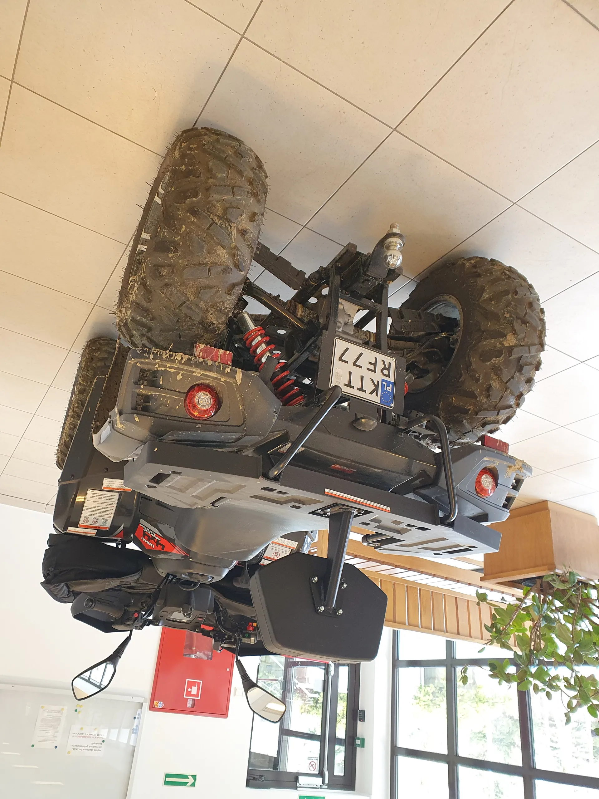 Czarny quad z brudnymi oponami, widoczna tablica rejestracyjna KTT RF77. W tle czerwona szafa gaśnicza, okna, zielony znak ewakuacyjny, donica z rośliną oraz podłoga z białych płytek. Quad znajduje się w pomieszczeniu, opony pokryte są błotem, widoczne lusterka boczne i czerwone elementy zawieszenia.