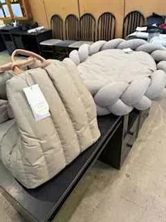 Duża, jasnoszara poduszka w stylu plecionym, obok niej beżowa pikowana kurtka z białą etykietą. Obiekty umieszczone na czarnej powierzchni. W tle widoczne drewniane krzesła z wygiętymi oparciami oraz betonowa ściana.