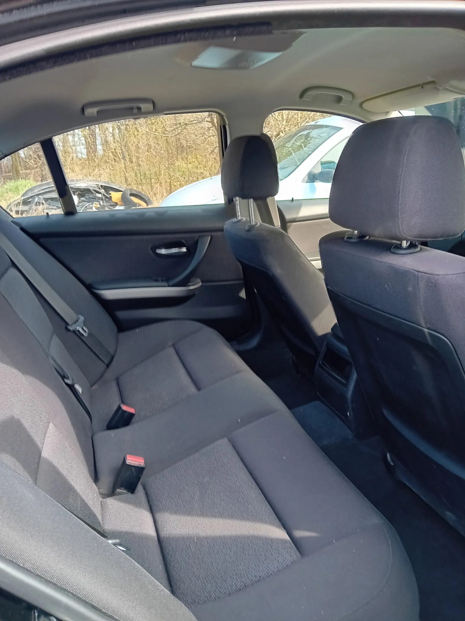 Wnętrze samochodu widziane z tyłu, przedstawiające czarne tylne siedzenia z pasami bezpieczeństwa, czarne przednie siedzenia z zagłówkami oraz fragment przedniej szyby i okien. Za szybą widoczny jest inny samochód oraz suche krzewy i drzewa na zewnątrz. Wnętrze jest oświetlone światłem słonecznym, rzucającym cienie na siedzenia.