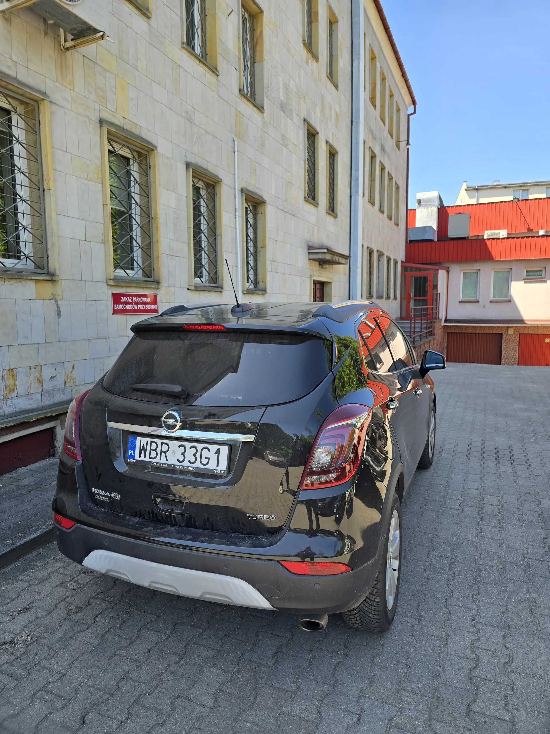 Czarny samochód marki Opel zaparkowany na brukowanej powierzchni. Tablica rejestracyjna WBR 33G1. Samochód znajduje się obok wielopiętrowego budynku z kamiennej elewacji i oknami z kratami. Na budynku czerwony znak z napisem ZAKAZ PARKOWANIA oraz SPOSOBEM PRZECIWNYM. W tle inne budynki z czerwonymi i beżowymi elewacjami. Niebo jest jasne i bezchmurne.