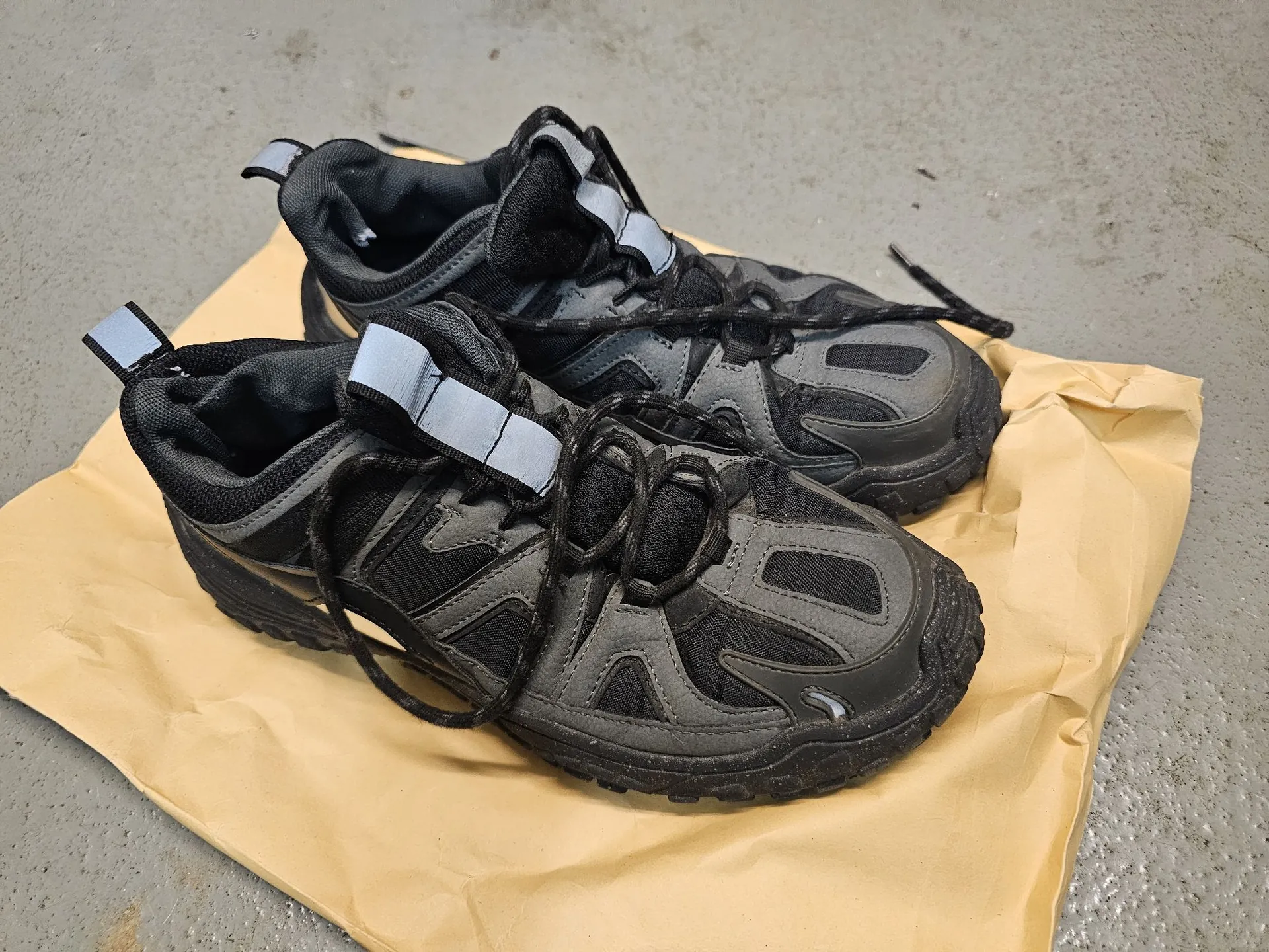 Para czarnych butów sportowych z szarymi elementami i odblaskowymi paskami przy językach, leżących na arkuszu brązowego papieru pakowego na szarym, betonowym podłożu.