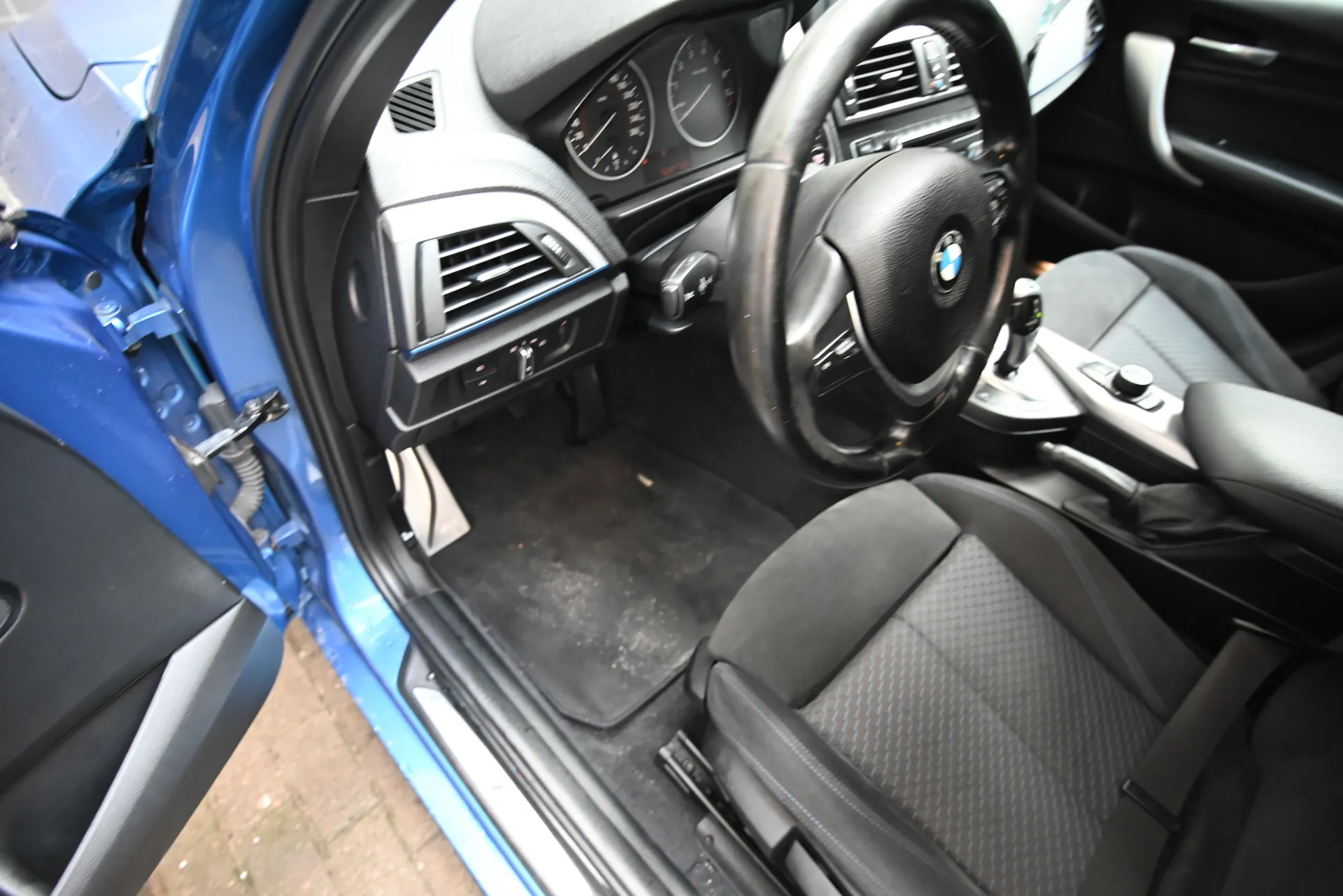 Wnętrze niebieskiego samochodu z otwartą drzwiami. Widoczna czarna kierownica z logo BMW, deska rozdzielcza z dwoma okrągłymi wskaźnikami, czarne siedzenia z geometrycznym wzorem, brudne dywany podłogowe oraz fragment niebieskiej obudowy drzwi z odsłoniętymi elementami.