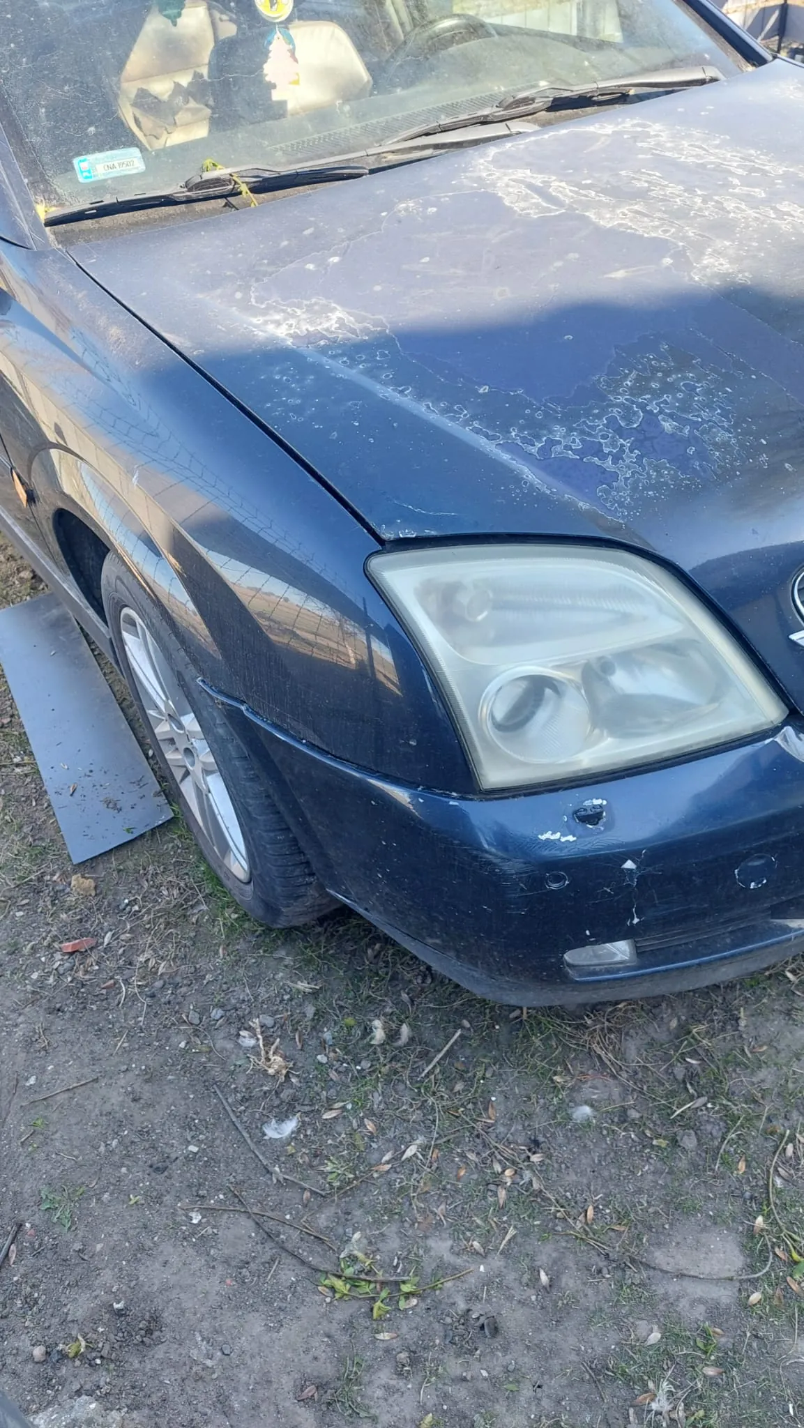 Ciemnoniebieski samochód widoczny od przodu, z widocznym maskiem pokrytym białymi plamami, reflektorem z dwoma żarówkami oraz uszkodzeniami na zderzaku. Na przedniej szybie naklejka z nieczytelnym tekstem. Samochód zaparkowany na brudnej ziemi z trawą i rozsypanymi liśćmi, pod przednim kołem metalowa płyta. Opona i felga częściowo widoczne.