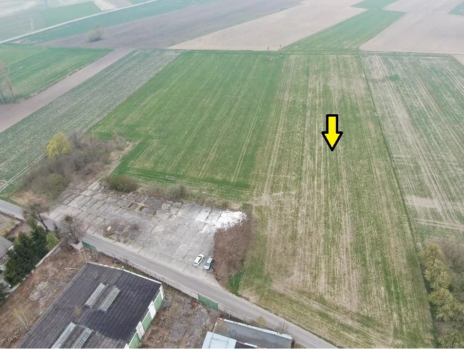 Zdjęcie lotnicze przedstawiające krajobraz rolniczy z polami o różnych odcieniach zieleni i brązu. W dolnej części obrazu widoczny jest betonowy plac z zaparkowanymi dwoma samochodami oraz fragment dachu dużego budynku przemysłowego. Na jednym z zielonych pól znajduje się żółta strzałka skierowana w dół.