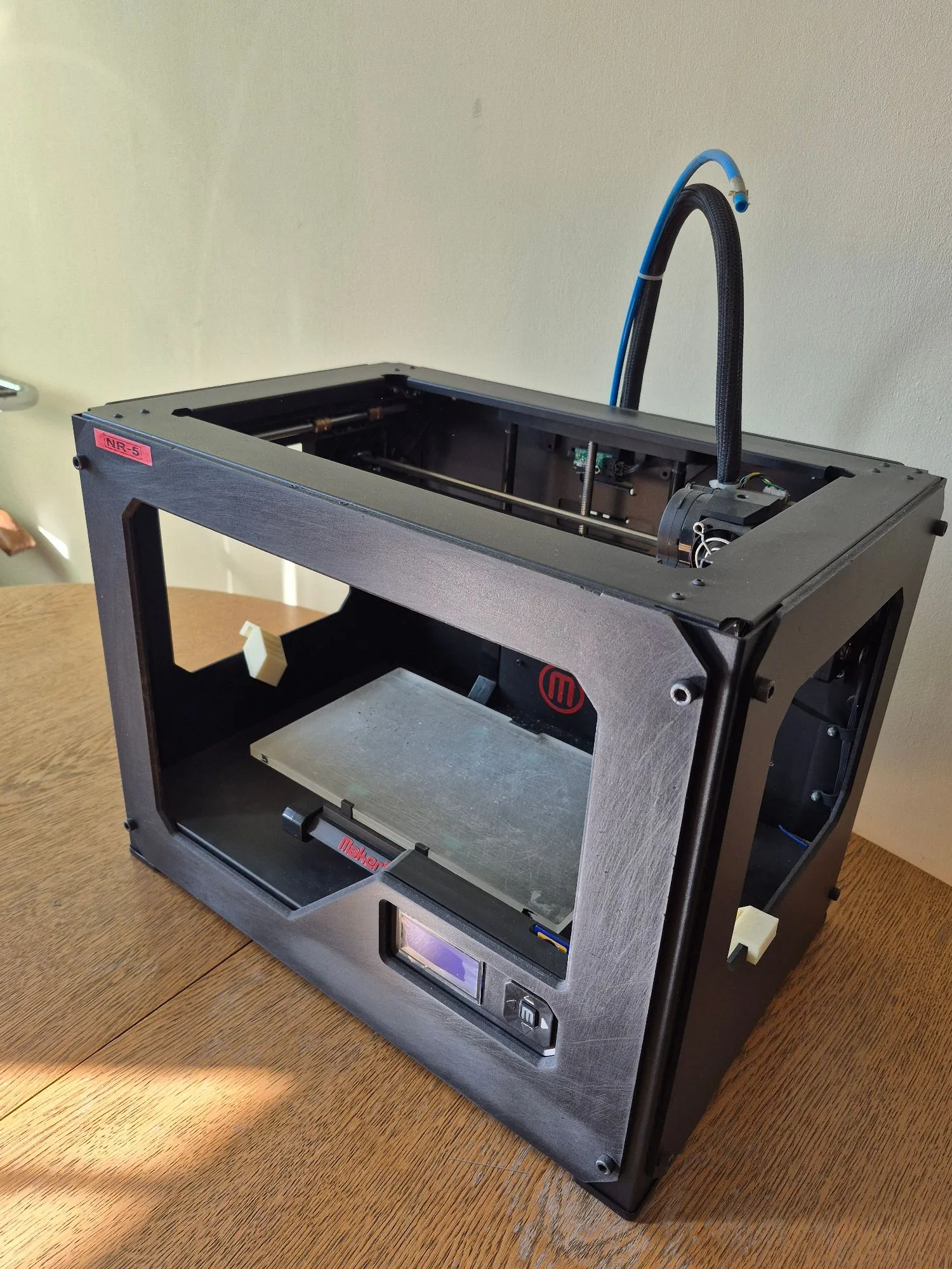 Czarna, zamknięta obudowa do drukarki 3D stojąca na drewnianym stole. Obudowa ma prostokątny kształt z dużym otworem z przodu i po bokach. Wewnątrz widoczna jest platforma robocza oraz mechanizmy drukarki. Na górze wystaje czarny przewód z niebieską rurką. W lewym górnym rogu obudowy znajduje się czerwona naklejka z napisem 'NR-5', a na przedniej ścianie logo 'm' w czerwonym okręgu oraz napis 'Maker'. Na dole z przodu widoczny jest mały ekran kontrolny i przełącznik.