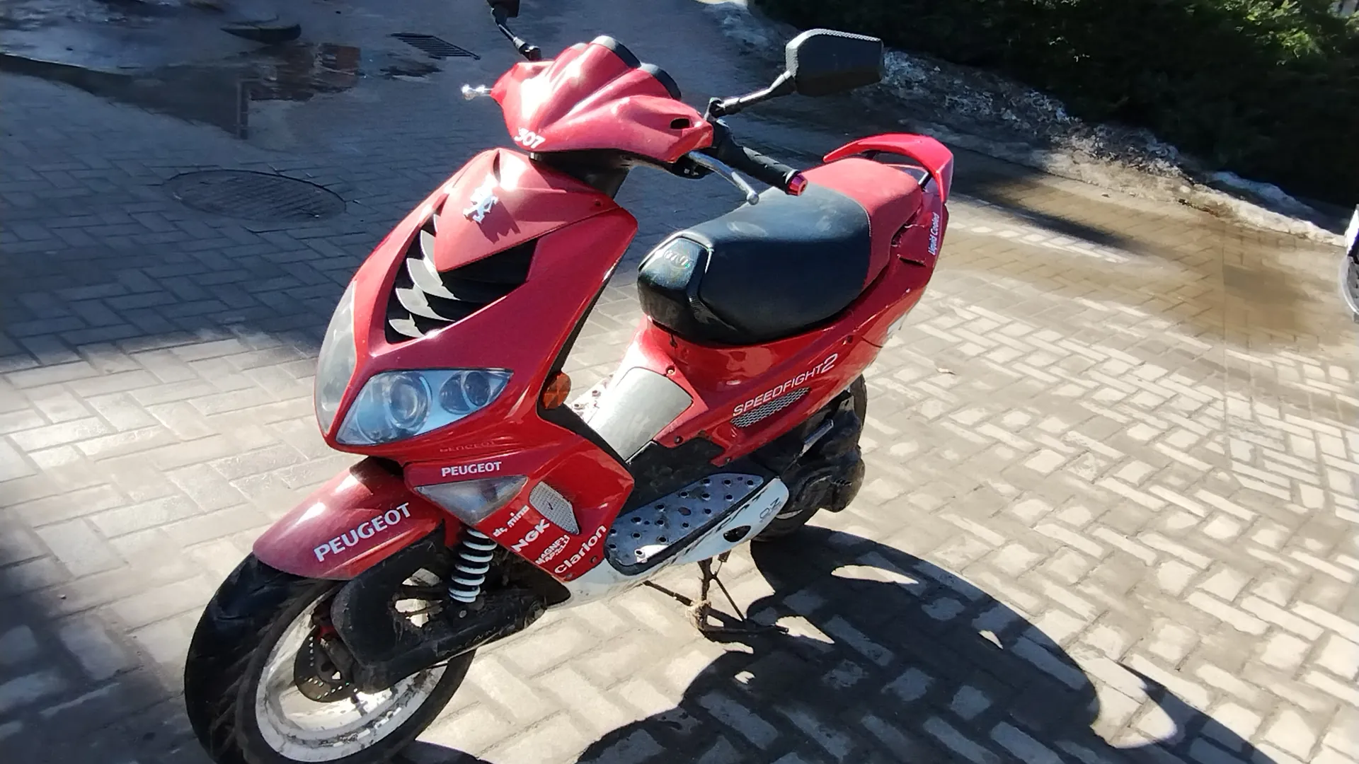 Czerwony skuter marki Peugeot model Speedfight 2 zaparkowany na brukowanej powierzchni. Na skuterze widoczne napisy: PEUGEOT na przodzie i boku, SPEEDFIGHT 2 na boku, oraz logotypy NGK i Clarion. Skuter posiada czarne siedzenie, lusterka boczne, reflektory i numer 307 na kierownicy. W tle brukowana ulica z kałużami i zieleń.