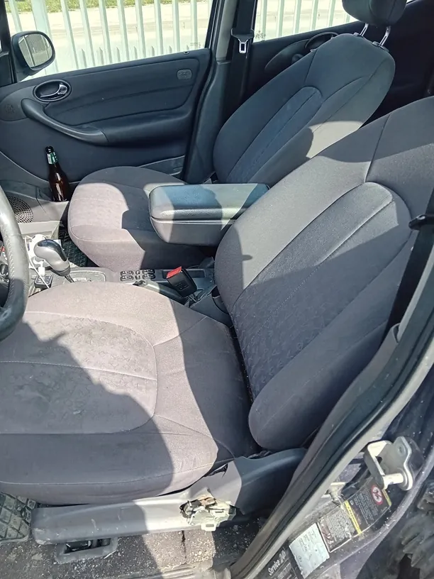 Wnętrze samochodu z widocznymi przodowymi siedzeniami tapicerowanymi szarym materiałem. Na siedzeniu kierowcy widoczne ślady zużycia. W otwartym drzwiu kierowcy znajduje się butelka. W centralnej konsoli widoczny dźwignia zmiany biegów oraz przyciski. Poza samochodem metalowe ogrodzenie. Kierownica po lewej stronie, co wskazuje na lewostronny napęd.