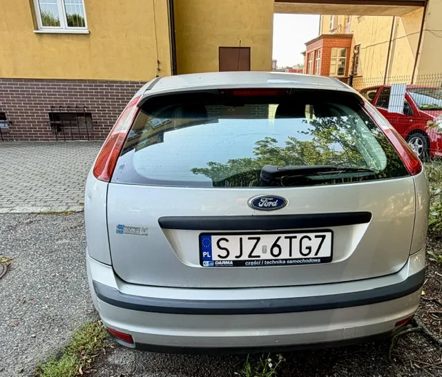 Srebrny samochód marki Ford widziany od tyłu, zaparkowany na kostce brukowej przed żółtym budynkiem. Na klapie bagażnika znajduje się logo Ford oraz polska tablica rejestracyjna o numerze SJZ 6TG7. Poniżej numeru rejestracyjnego widnieje napis 'DARMA części i technika samochodowa'.