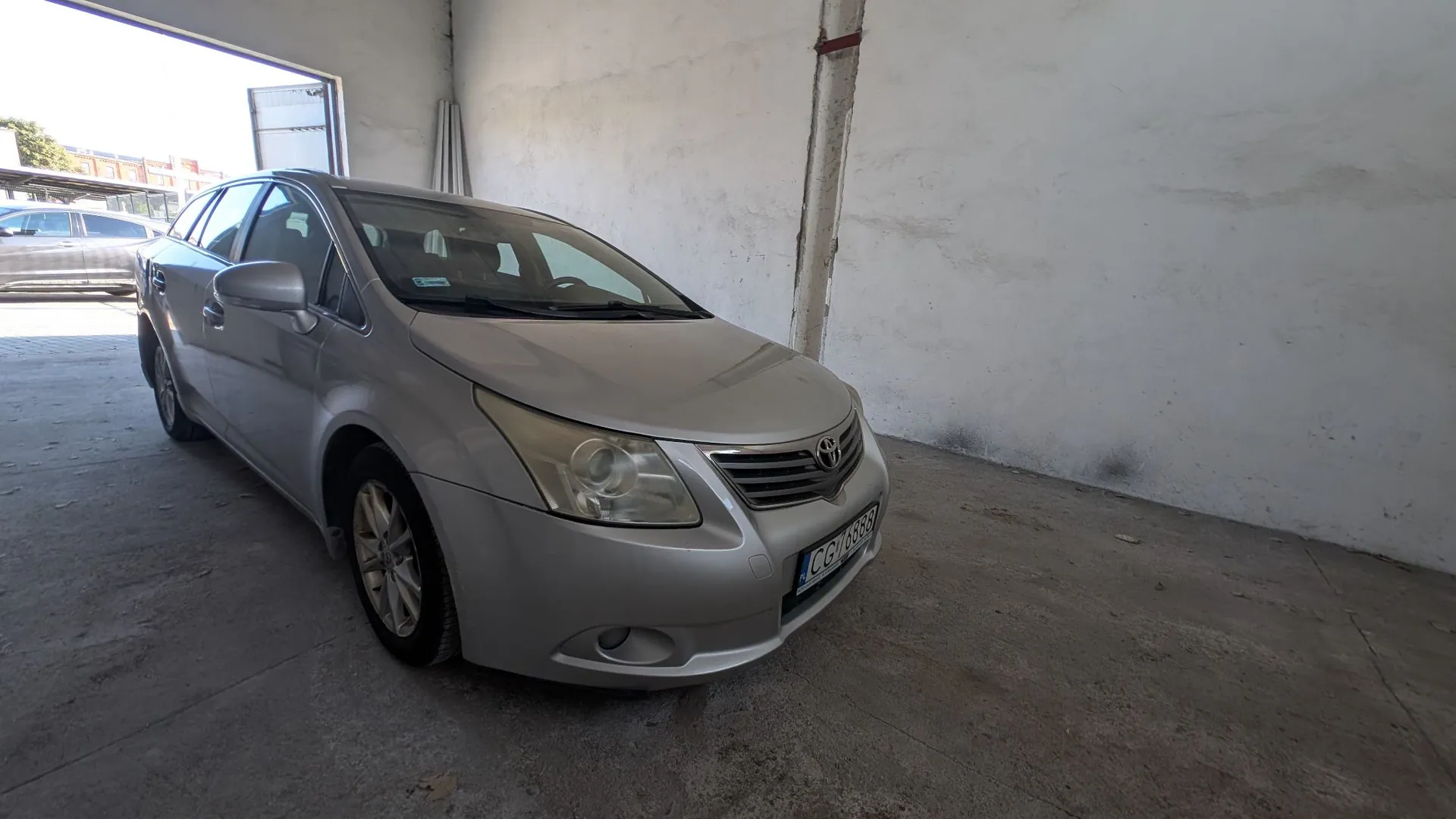 Srebrny samochód osobowy marki Toyota zaparkowany wewnątrz jasnego garażu lub hali z betonową podłogą i białymi ścianami. Pojazd jest ustawiony pod kątem, widoczny z przodu i z boku. Na przednim zderzaku znajduje się tablica rejestracyjna o numerze CGY 1888.