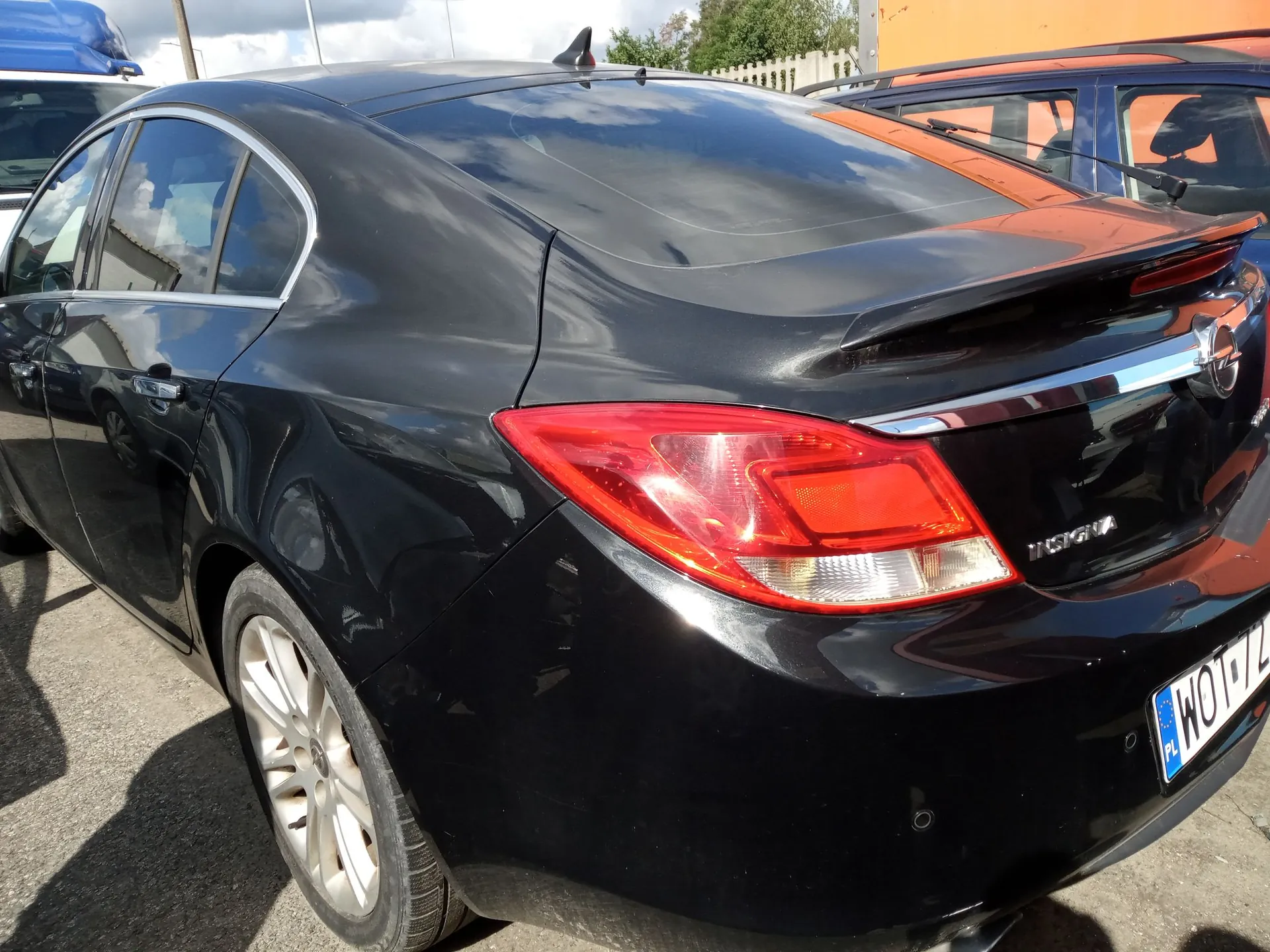 Tylna część czarnego samochodu marki Opel Insignia, widoczna z perspektywy trzech czwartych. Na klapie bagażnika znajduje się napis 'INSIGNIA' oraz logo marki. Widoczna jest prawa lampa tylna oraz fragment tablicy rejestracyjnej zaczynający się od liter 'WOT'. W tle widać fragment innego, niebieskiego samochodu oraz pomarańczową ścianę budynku.