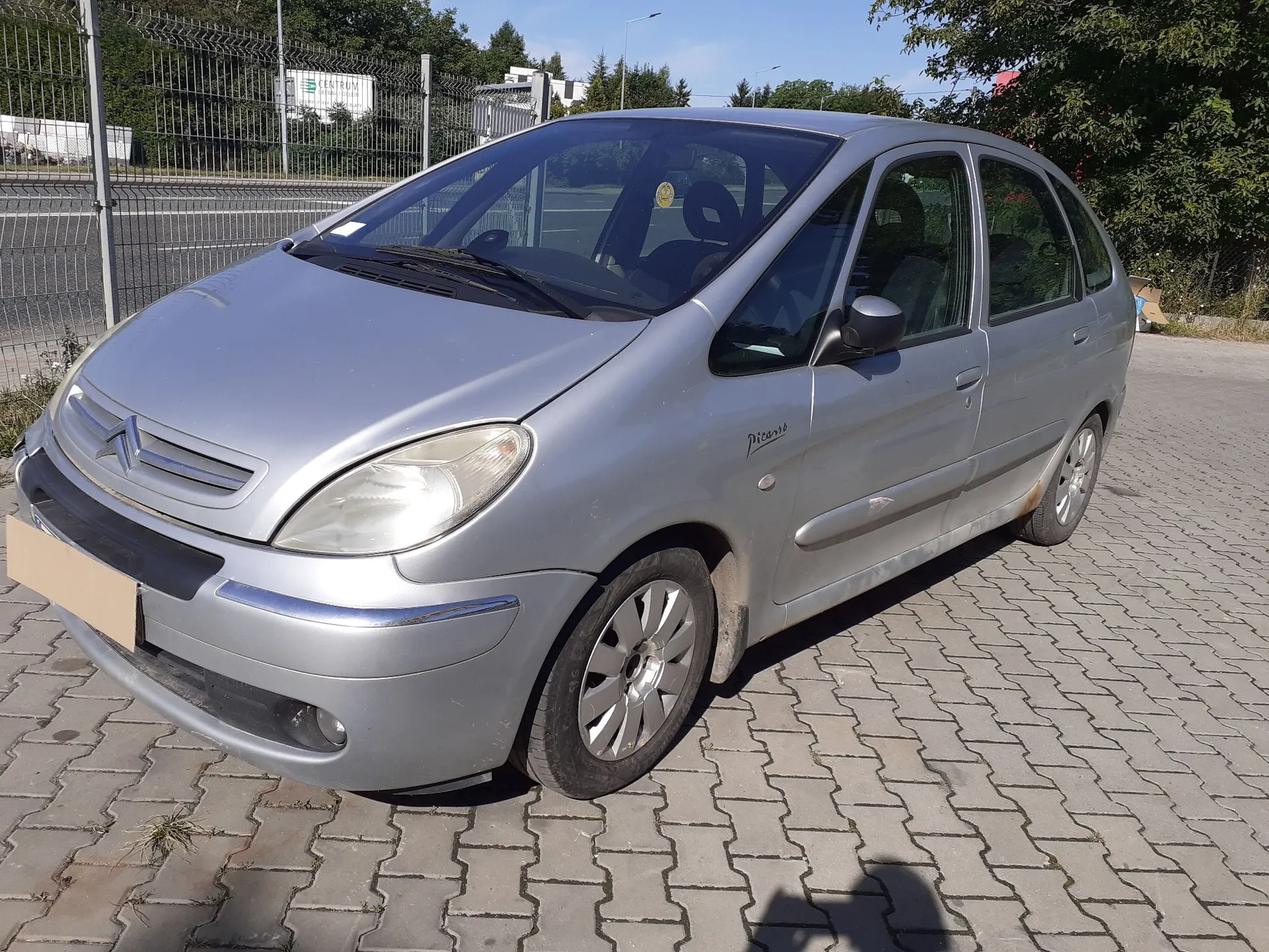 Srebrny samochód marki Citroën Picasso zaparkowany na kostce brukowej. Pojazd jest widoczny z przodu i z boku, pod kątem. W tle znajduje się metalowy płot, drzewa oraz fragmenty budynków pod jasnym, niebieskim niebem.