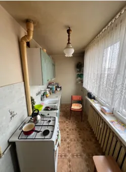 Kuchnia z białą kuchenką gazową z czerwonym garnkiem i czarną patelnią na blacie. Po lewej stronie zlewozmywak z naczyniami, nad nim szafki. Na ścianie biegnie żółty rurociąg. Przy oknie z koronkową zasłoną znajduje się grzejnik i ławka. W środku pomieszczenia krzesło, podłoga z wzorzystą płytką. Sufit z wiszącą lampą.