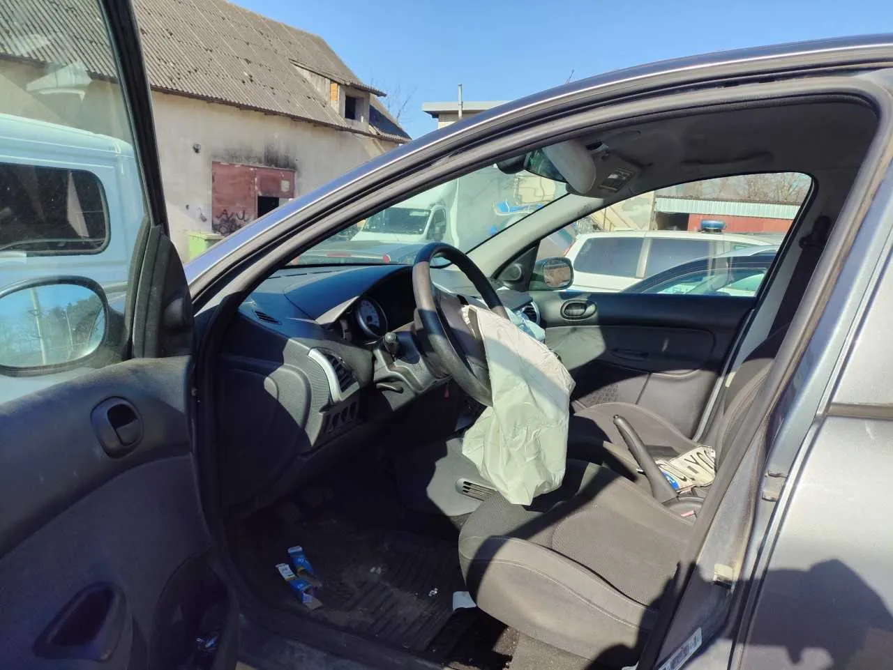 Wnętrze samochodu z otwartą drzwiami po stronie kierowcy. Widoczna deska rozdzielcza, kierownica oraz rozwinięta poduszka powietrzna po stronie kierowcy. Siedzenia są szare, na podłodze leżą niebieskie opakowania. W tle budynki, w tym jeden z uszkodzonym dachem i czerwoną bramą, oraz inne zaparkowane pojazdy. Niebo jest bezchmurne.