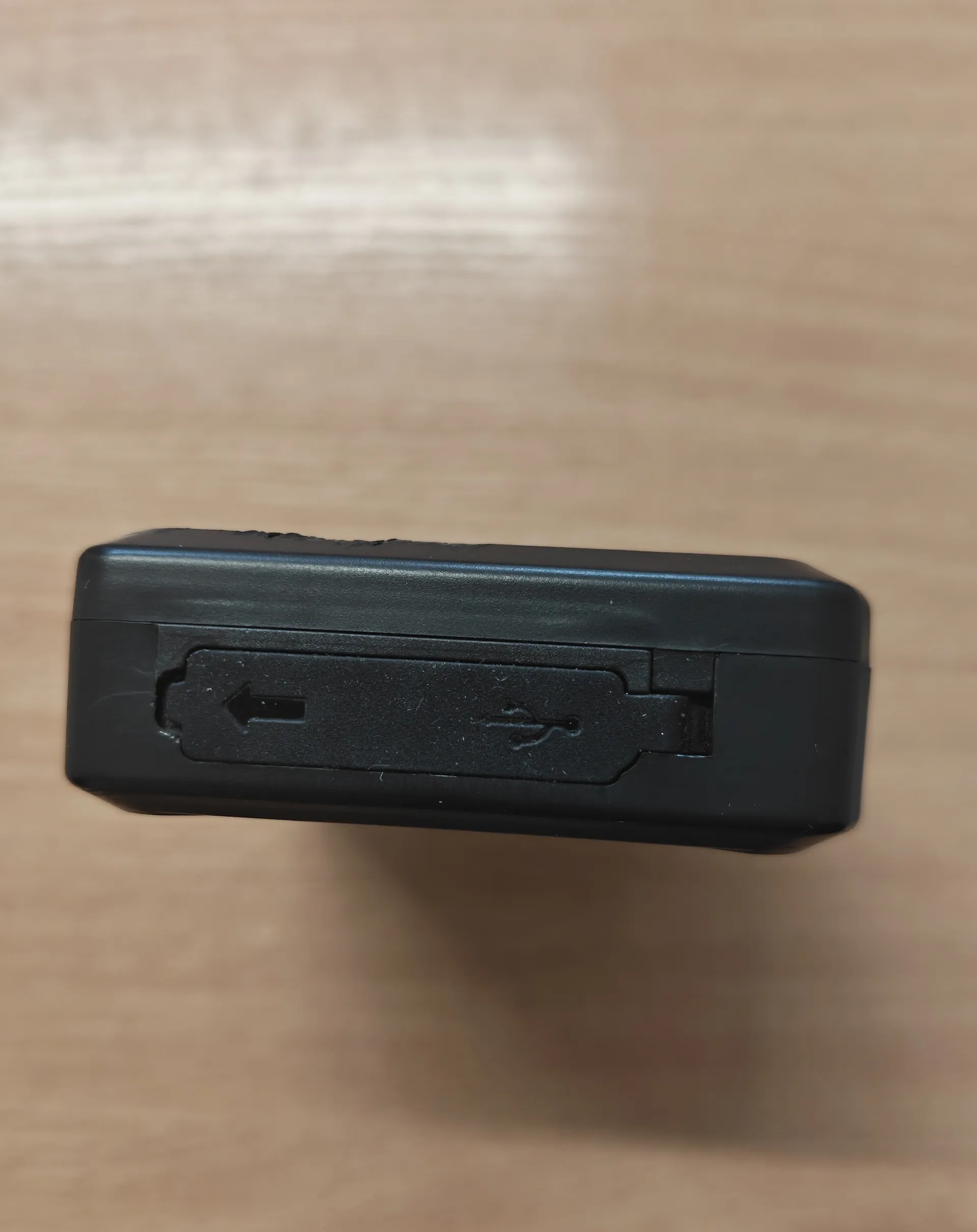 Zbliżenie na czarną, plastikową obudowę urządzenia elektronicznego leżącego na drewnianym blacie. Widoczny jest port ładowania lub przesyłu danych z symbolem USB oraz strzałką wskazującą kierunek wkładania wtyczki.
