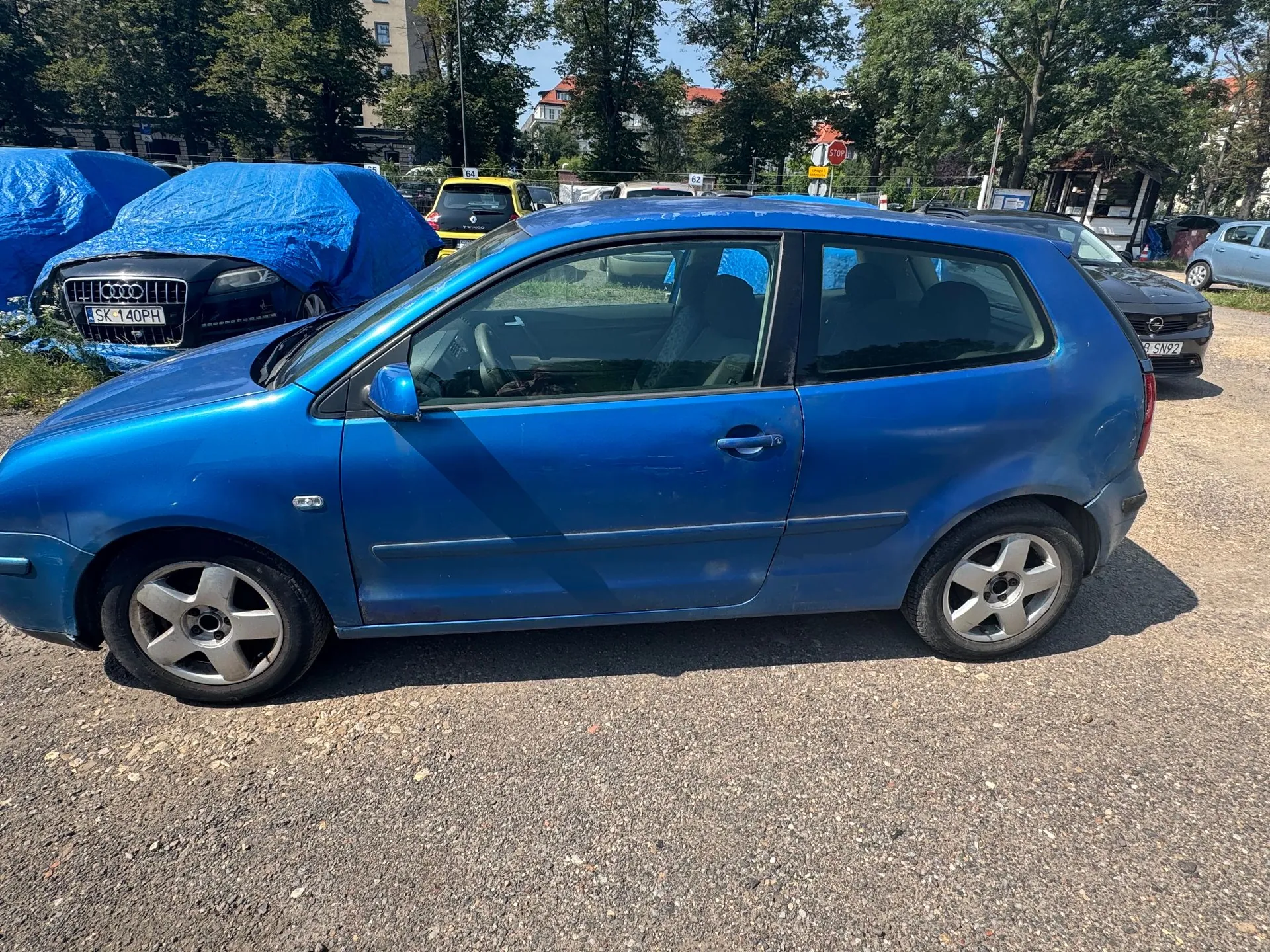 Niebieski samochód osobowy typu hatchback zaparkowany bokiem na żwirowym placu. W tle widoczne są inne pojazdy, w tym samochód przykryty niebieską plandeką z widoczną tablicą rejestracyjną SK 140PH oraz żółty samochód. W oddali znajdują się drzewa i budynki.
