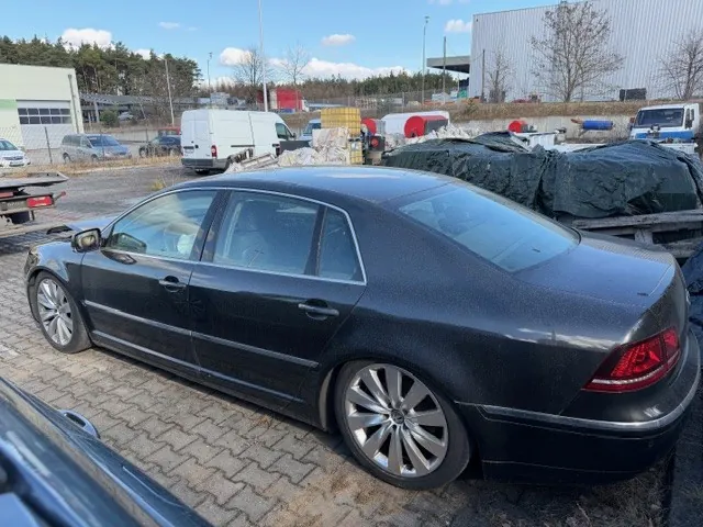 Czarny samochód osobowy typu sedan zaparkowany na brukowanej powierzchni. W tle widoczne są inne pojazdy, w tym biała furgonetka i ciężarówka, przemysłowe budynki, stosy materiałów przykryte czarną folią oraz drzewa. Niebo jest częściowo zachmurzone. Samochód znajduje się w centralnej części kadru, z widocznymi srebrnymi felgami i czerwonymi światłami tylnymi.