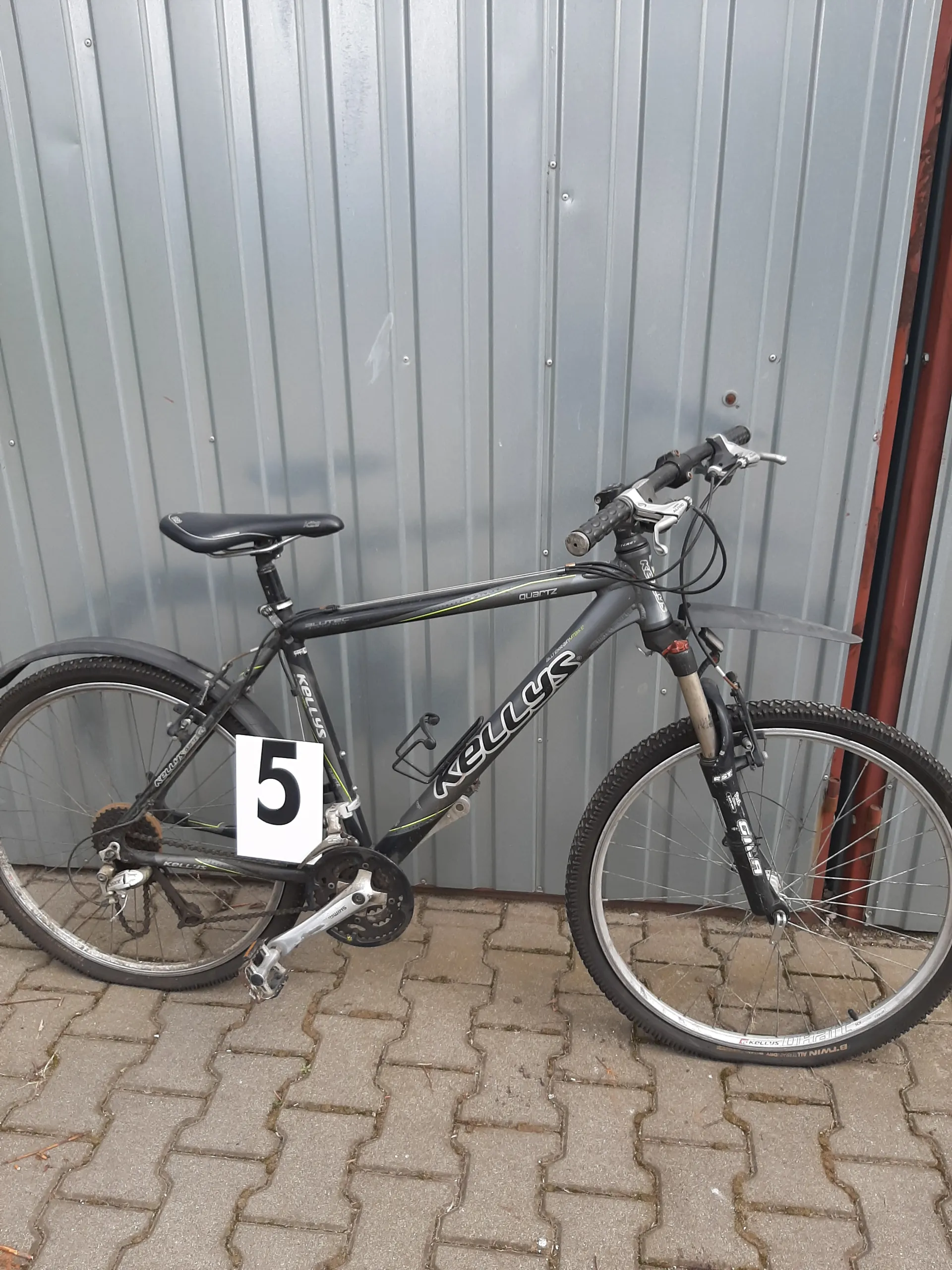 Czarny rower górski marki KELLYS z białym znakiem z czarną cyfrą 5 na ramie. Rower oparty o szarą metalową bramę garażową z poziomymi listwami i brukowaną powierzchnię z kamiennych płyt. Widoczne elementy: kierownica z hamulcami, siedzisko, koła z oponami, łańcuch i przerzutki.