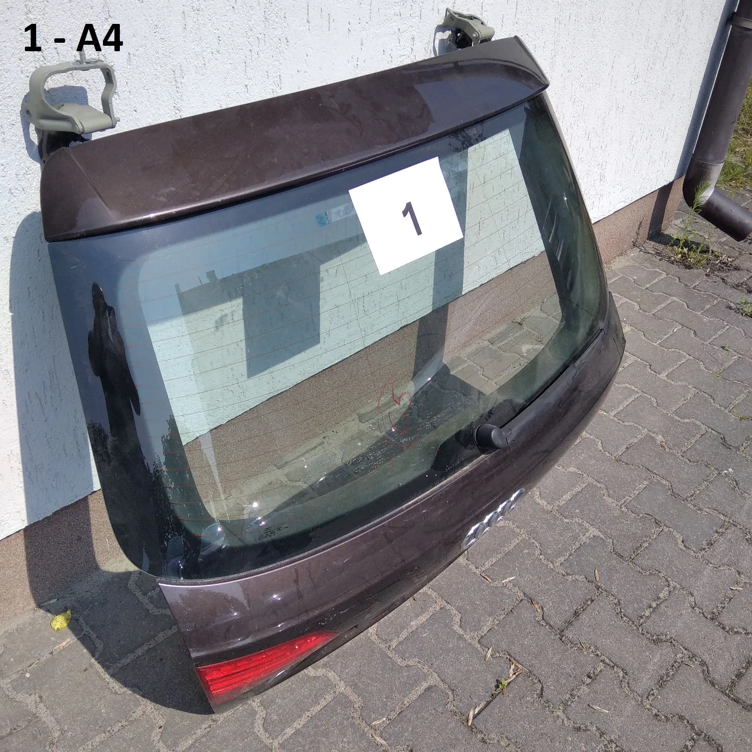 Zdjęcie tylnej klapy bagażnika z szybą z samochodu marki Audi A4 w kolorze ciemnobrązowym. Klapa oparta jest o białą ścianę na kostce brukowej. Na szybie znajduje się biała kartka z czarną cyfrą '1'. W lewym górnym rogu obrazu widnieje napis '1 - A4'.