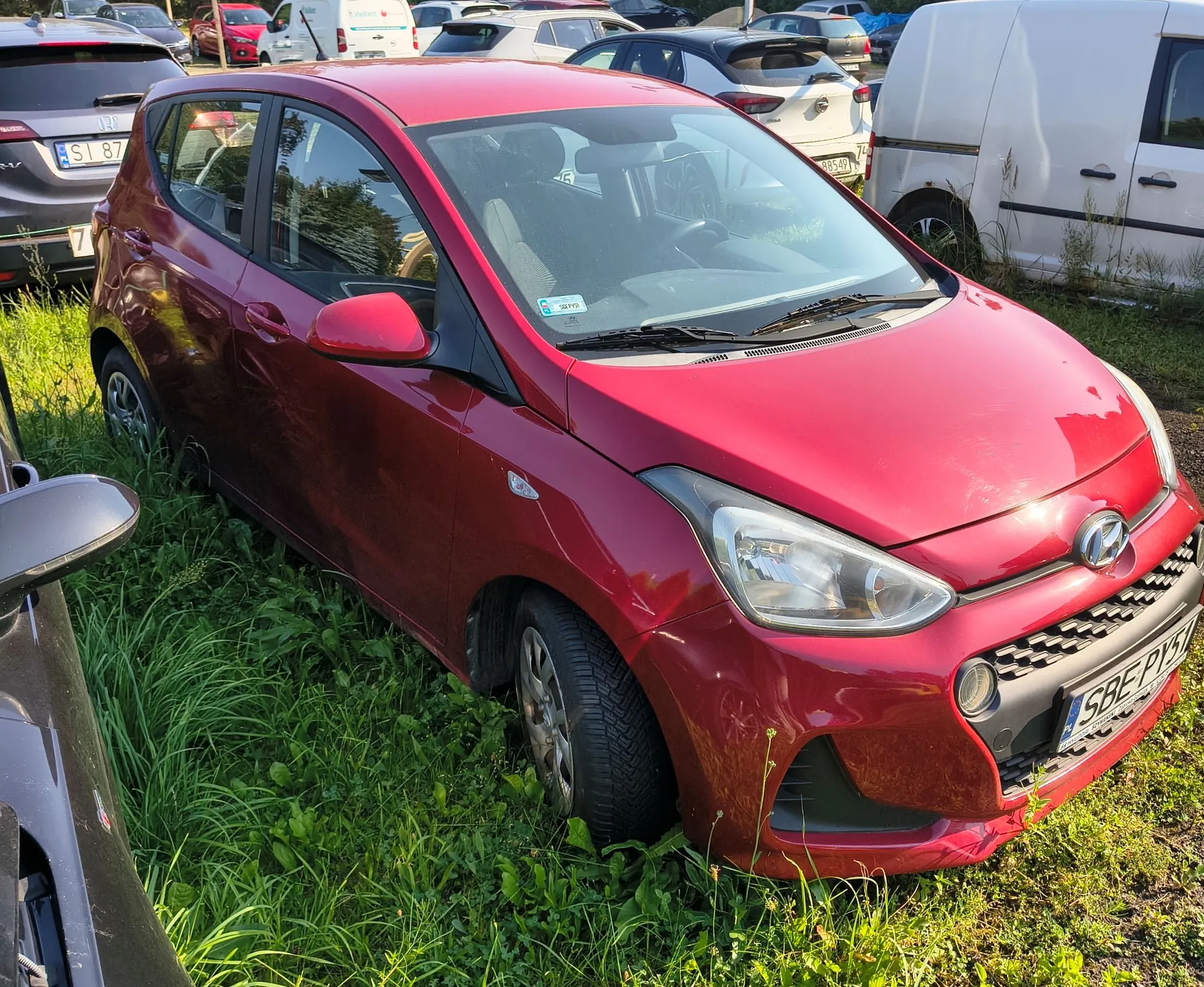 Czerwony samochód osobowy marki Hyundai zaparkowany na trawie. Pojazd jest widoczny z przodu i z boku. W tle znajdują się inne samochody, w tym biały furgon oraz srebrny samochód z tablicą rejestracyjną SL 87. Na przednim zderzaku czerwonego auta widoczna jest tablica rejestracyjna SBE PY... (częściowo zasłonięta).