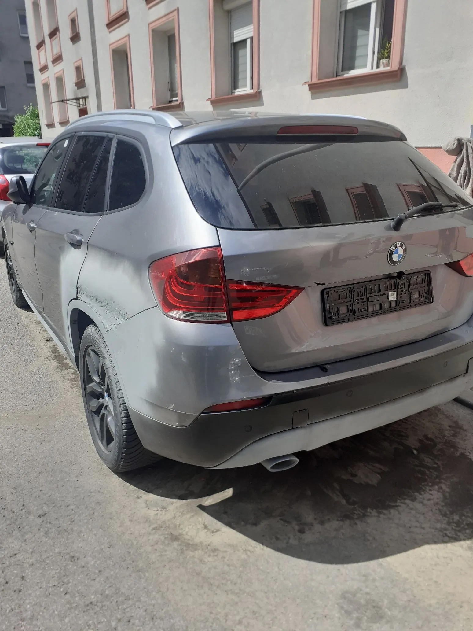 Srebrny samochód marki BMW typu SUV zaparkowany przy budynku. Pojazd ma widoczne uszkodzenia karoserii w postaci wgnieceń i zarysowań na lewym tylnym błotniku oraz tylnych drzwiach. Samochód nie posiada tablic rejestracyjnych.