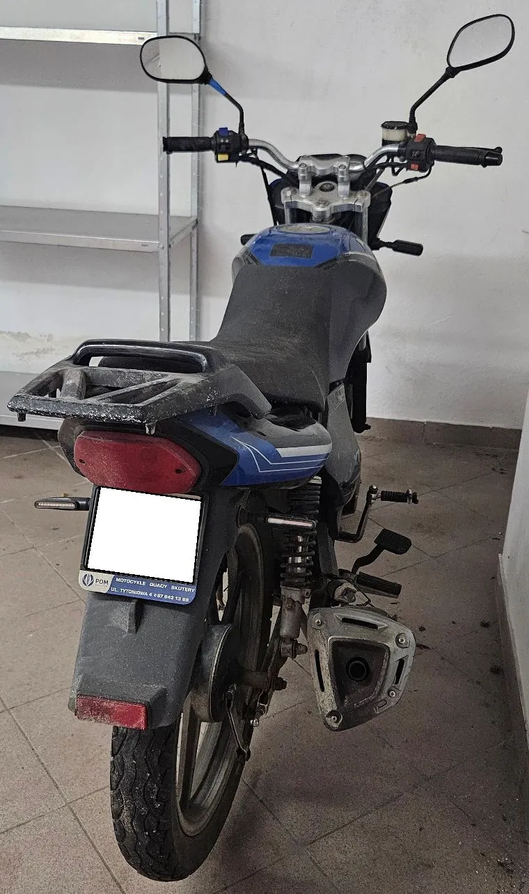 Widok z tyłu na niebiesko-czarny motocykl zaparkowany w pomieszczeniu na szarym kafelkowym podłożu. Motocykl posiada czarne siedzenie, tylny bagażnik oraz lusterka. Pod tylną lampą znajduje się biała tablica rejestracyjna oraz ramka z napisem: 'POM MOTORCYKLE QUADY BATERY UL. TYTONOWA 1 + 67 843 12 85'. W tle widoczny jest metalowy regał.