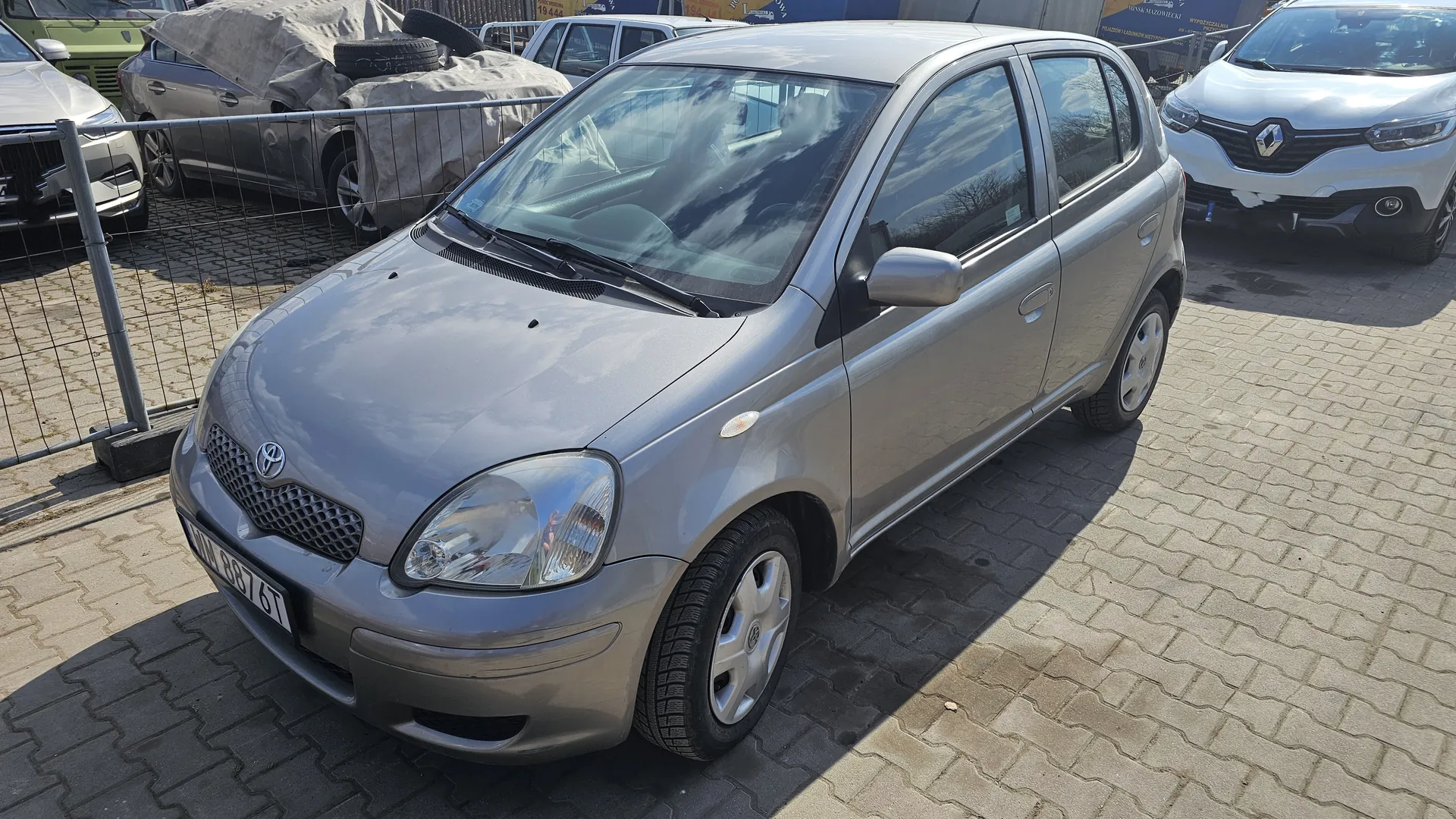 Srebrny samochód osobowy marki Toyota z tablicą rejestracyjną WU 887 GT zaparkowany na brukowanej powierzchni. W tle widoczne są inne pojazdy, w tym zakryty tkaniną samochód, biały Renault oraz metalowe ogrodzenie. Na niebie znajdują się chmury, a całość znajduje się na otwartym placu parkingowym.
