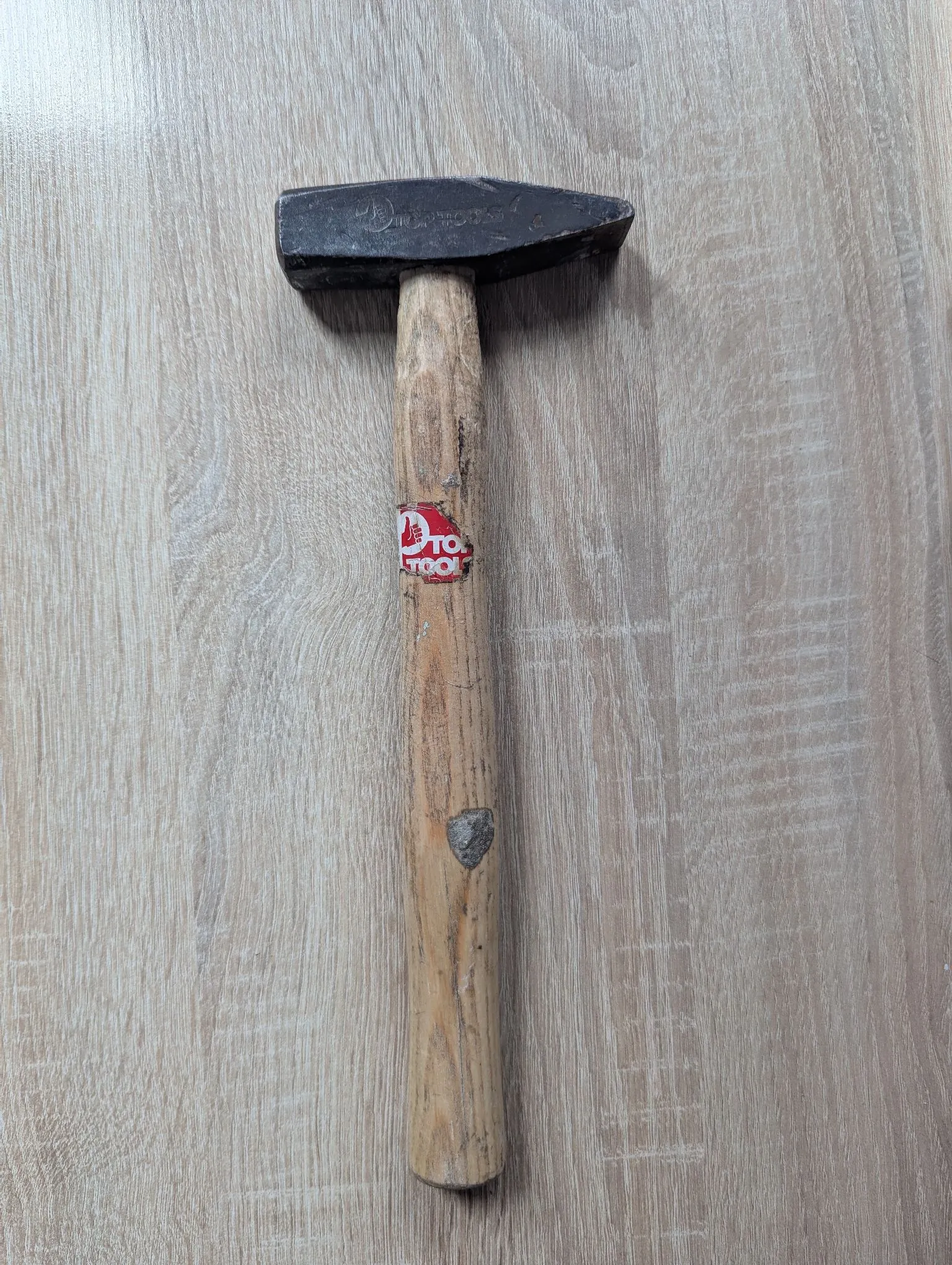 Młotek z metalową główką i drewnianym trzonkiem, leżący pionowo na jasnym, drewnianym blacie. Na trzonku znajduje się czerwono-biała naklejka z napisem 'TOOL'.