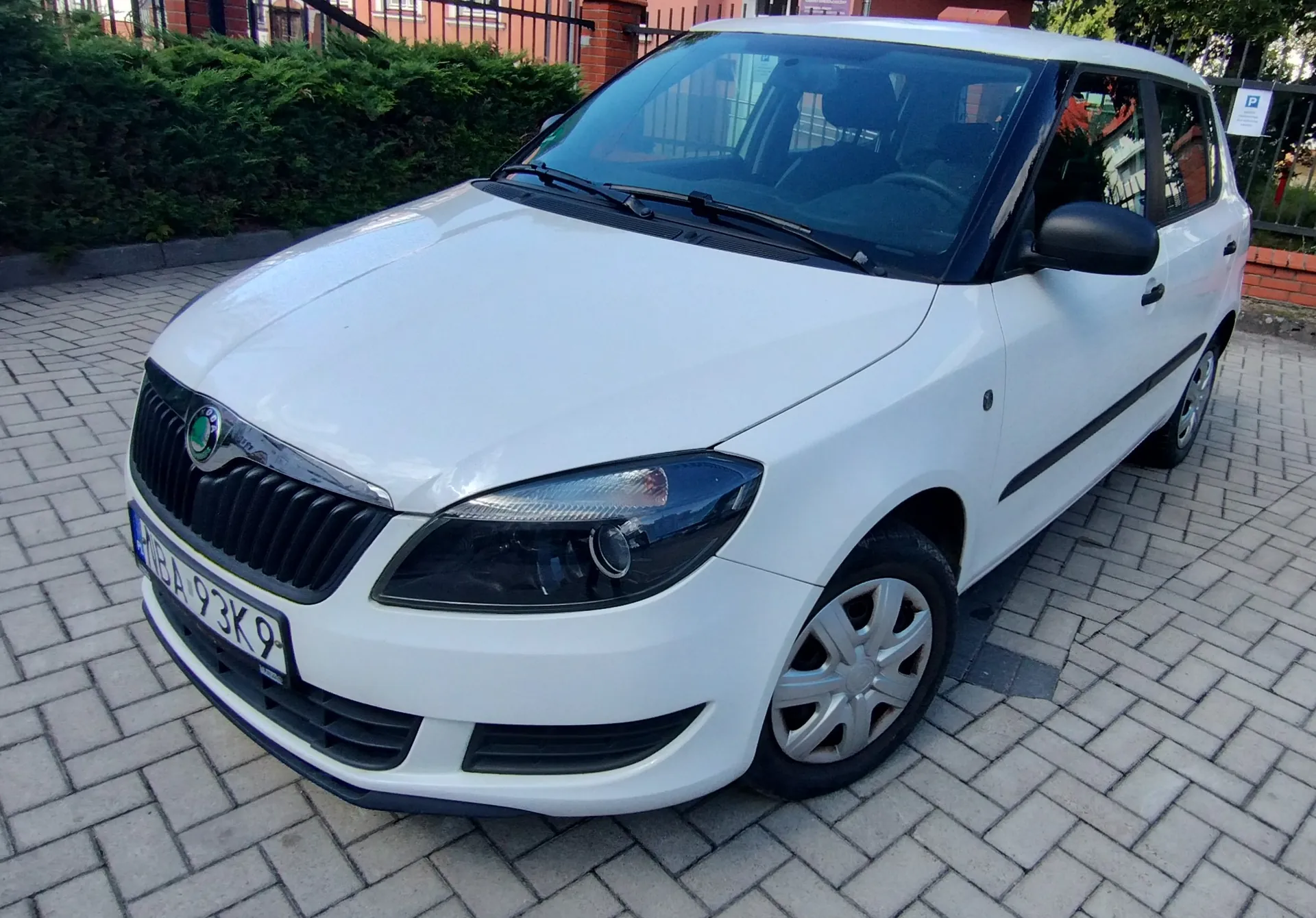 Biały samochód osobowy marki Skoda zaparkowany na kostce brukowej. Pojazd jest widoczny z przodu pod kątem. Na samochodzie widoczna jest polska tablica rejestracyjna o numerze BA 93K9.