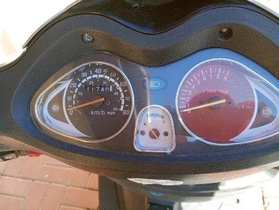 Zbliżenie na deski rozdzielcze motocykla z dwoma głównymi wskaźnikami. Po lewej stronie znajduje się prędkościomierz z zakresem do 80 km/h, wskazujący 0 km/h, z licznikiem przebiegu 11740,1 km. Po prawej stronie czerwony tachometr z zakresem do 14 000 obr./min, wskazujący około 2000 obr./min. W centrum mały wskaźnik paliwa. Tło przedstawia ceglane płytki chodnika. Całość umieszczona na czarnym panelu z niebieskimi i czerwonymi elementami.