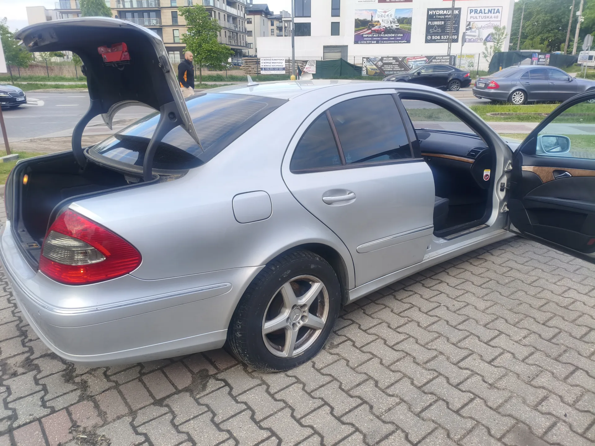 Srebrny samochód osobowy typu sedan z otwartym bagażnikiem i otwartą przednią drzwiami pasażerskimi. Samochód stoi na brukowanej powierzchni. W tle widoczne są budynki mieszkalne, tablice reklamowe z napisami 'HYDRAULIK', 'PRALNIA PŁATNA 48', 'PLUMBER' oraz inne zaparkowane pojazdy. Na tle stoi mężczyzna w czarnej kurtce i beżowych spodniach. Opony samochodu mają metalowe felgi, a wnętrze posiada czarne tapicerki z brązowymi wstawkami.