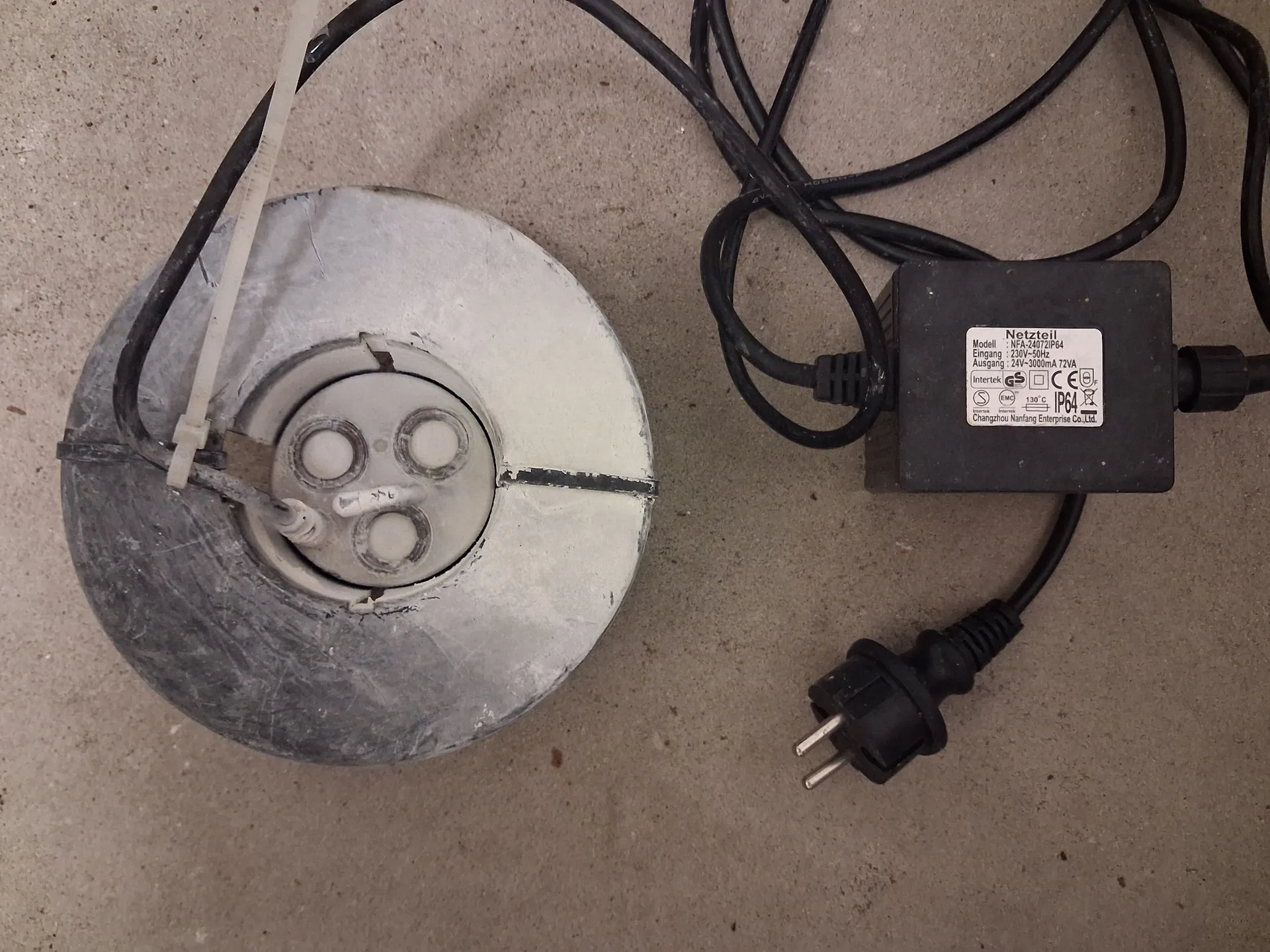 Zdjęcie przedstawiające urządzenie elektryczne składające się z okrągłej, metalowej obudowy w kolorze srebrnym oraz czarnego zasilacza z wtyczką sieciową. Zasilacz posiada białe napisy, w tym słowo 'Netzteil' oraz dane techniczne: 'Modell: HL-100100254', 'Eingang: 230V~50Hz', 'Ausgang: 12V=1000mA/12VA'. Urządzenia leżą na szarej, chropowatej powierzchni.