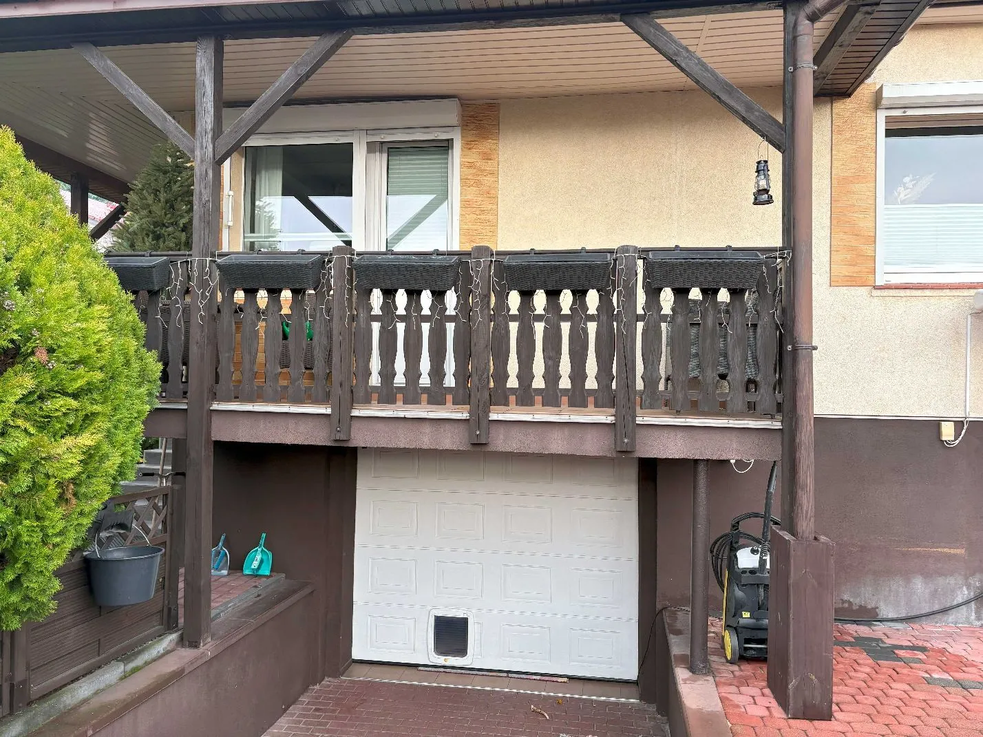 Zdjęcie przedstawiające fragment domu z tarasem na piętrze i garażem poniżej. Taras ma ciemnobrązowe drewniane balustrady z ozdobnymi tralkami i jest przykryty zadaszeniem wspartym na drewnianych słupach. Pod tarasem znajduje się biała, segmentowa brama garażowa. Po lewej stronie widoczny jest gęsty, zielony krzew, a obok niego szary kubeł. Po prawej stronie, przy ścianie garażu, stoi żółto-czarna myjka ciśnieniowa. Ściana domu jest w kolorze beżowym z brązowym pasem u dołu.
