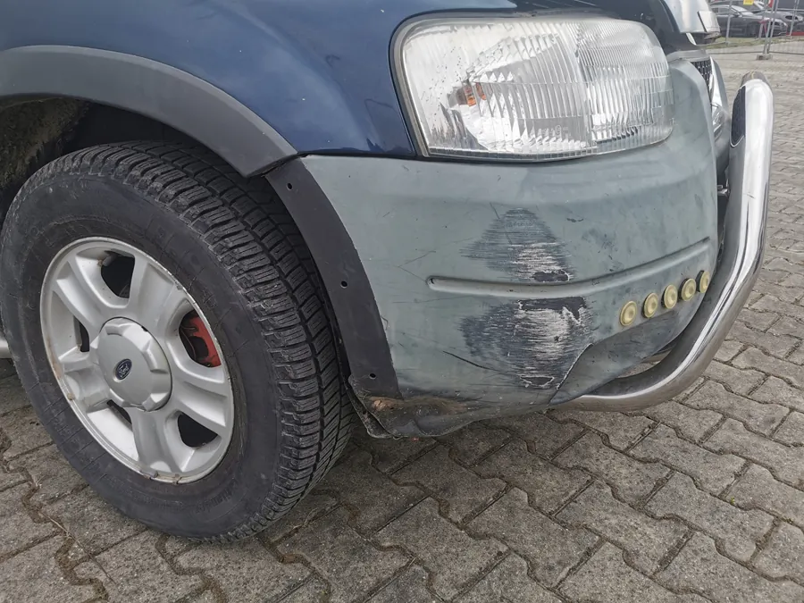 Zbliżenie przedniej lewej części niebieskiego samochodu. Widoczny uszkodzony zderzak z odsłoniętą warstwą podkładu, reflektor, srebrny bullbar z pięcioma okrągłymi elementami, koło z srebrną felgą i oponą z widocznym logo Ford. Samochód stoi na brukowanej powierzchni. Na zderzaku widoczne są zadrapania i uszkodzenia lakieru.