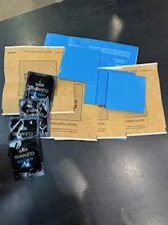 Na ciemnej powierzchni leżą brązowe kartonowe pudełka, niebieskie arkusze papieru oraz czarne opakowania z częściowo widocznym tekstem. Tekst na czarnych opakowaniach jest nieczytelny z powodu orientacji lub rozmiaru.