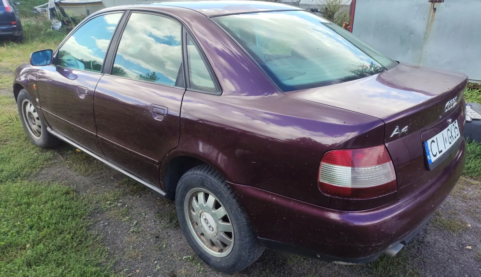 Purpurowy samochód osobowy marki Audi A4 zaparkowany na trawiasto-żwirowym podłożu. Widok z tyłu i z boku pojazdu. Na klapie bagażnika widnieje oznaczenie modelu 'A4' oraz tablica rejestracyjna o numerze 'CL V GK35'.