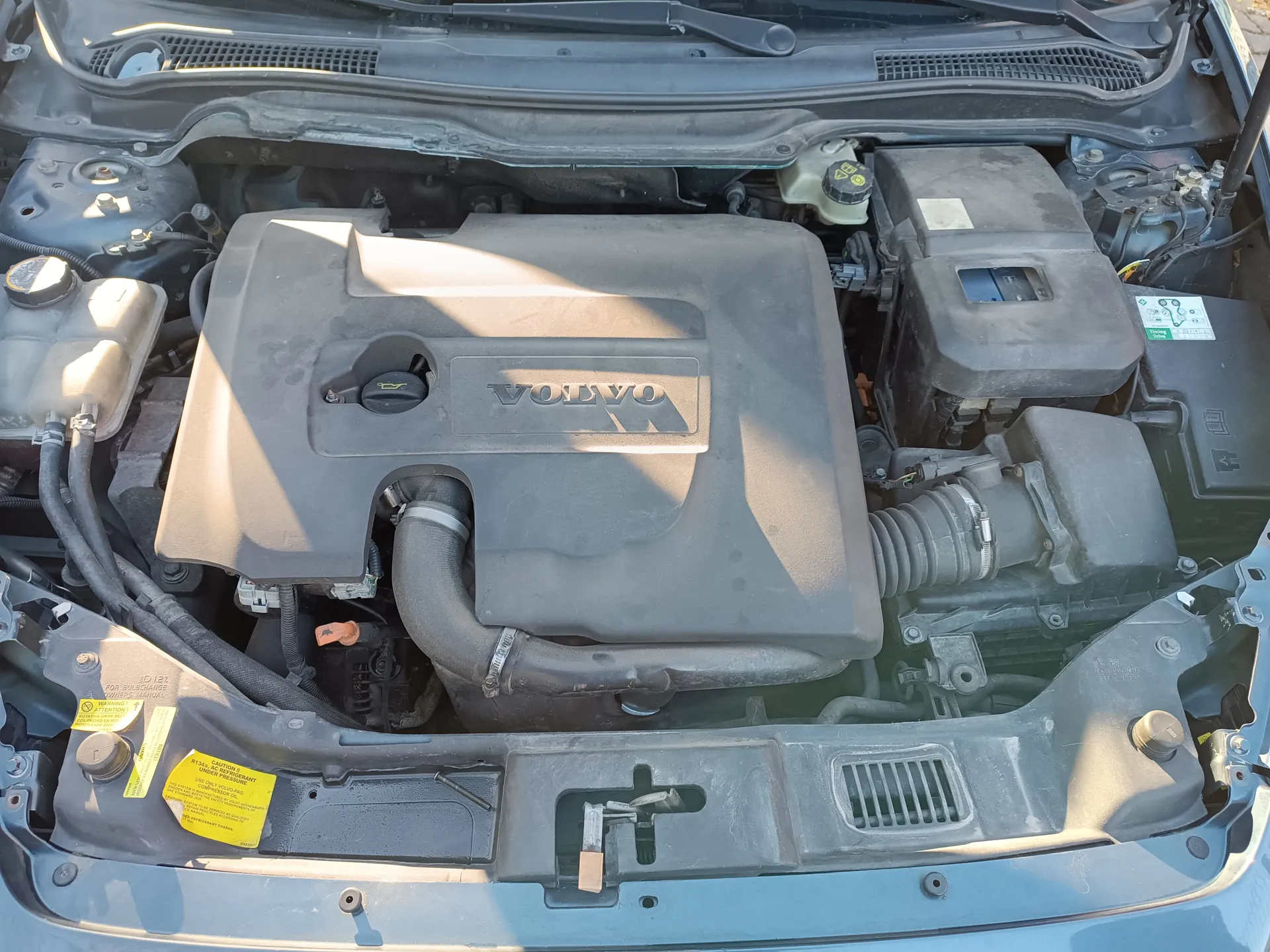 Widok otwartej maski samochodu marki Volvo z widocznym silnikiem. Na pokrywie silnika widnieje napis "VOLVO". Wokół znajdują się różne elementy silnika, w tym zbiorniki płynów, przewody i żółte etykiety ostrzegawcze z tekstem. Po prawej stronie widoczny jest czarny pojemnik z niebieskim okienkiem oraz bateria. Na dole obrazu znajduje się fragment przedniej części nadwozia samochodu.