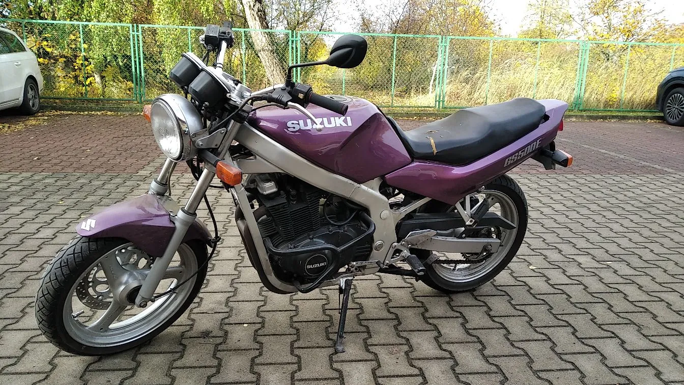 Fioletowy motocykl marki Suzuki zaparkowany na kostce brukowej. Motocykl jest ustawiony bokiem, z przodu widoczny jest okrągły reflektor, a na baku napis 'SUZUKI'. W tle znajduje się zielony płot z siatki oraz drzewa z jesiennymi liśćmi. Po lewej stronie widoczny jest fragment białego samochodu, a po prawej fragment czarnego samochodu.