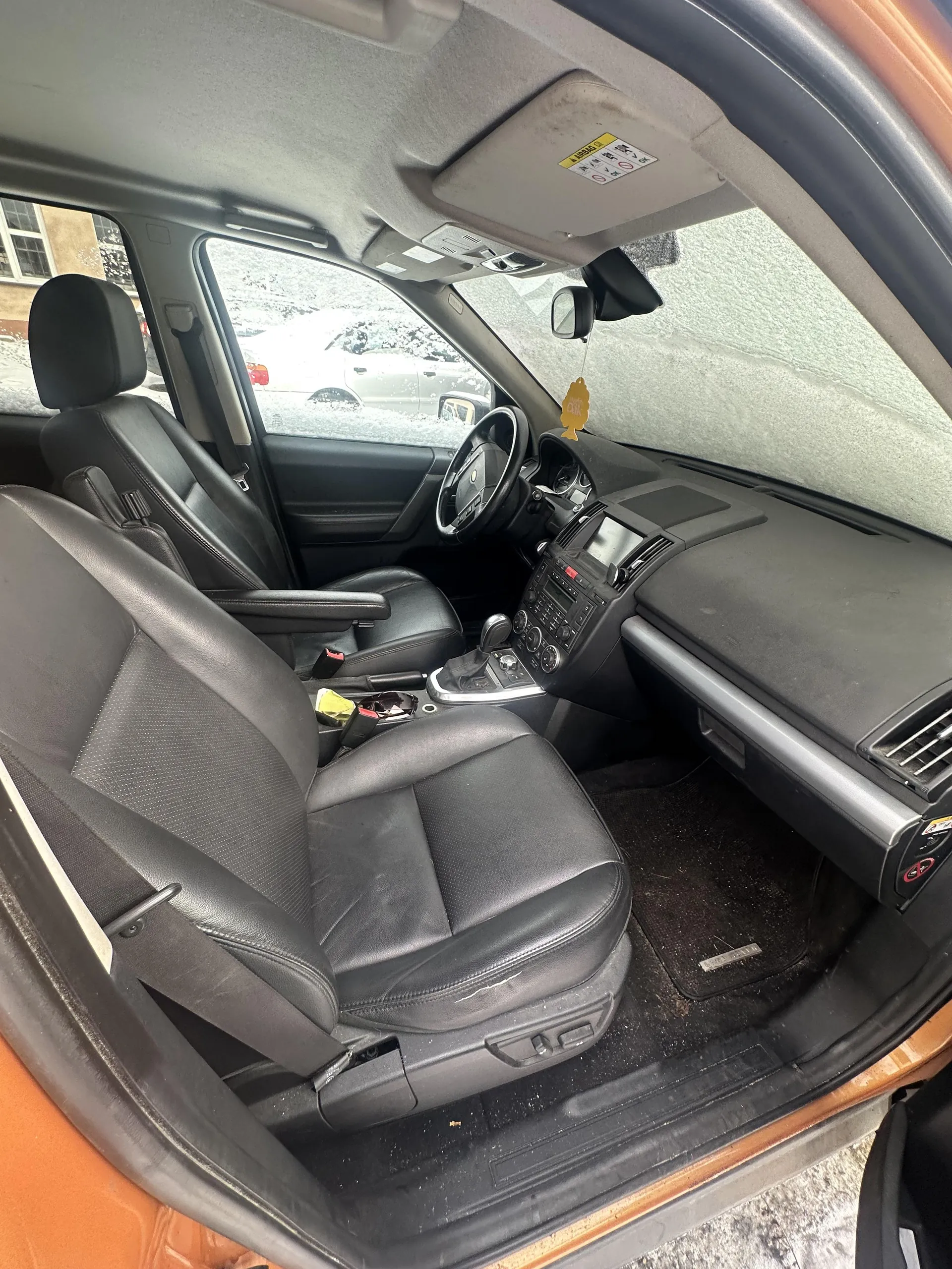 Wnętrze samochodu z czarnymi skórzany fotelami, kierownicą, deska rozdzielcza z radioodtwarzaczem i klimatyzacją. Na daszku przeciwsłonecznym widoczne ostrzeżenie z napisem 'A AIRBAG' i symbolami. W lusterku wstecznym wiszący żółty odświeżacz powietrza. Przez przednią szybę widać zasneżony teren i inne zaparkowane pojazdy. Drzwi pasażerskie są otwarte, ujawniając pomarańczowy kolor nadwozia. Podłoga pokryta czarnymi dywanikami.