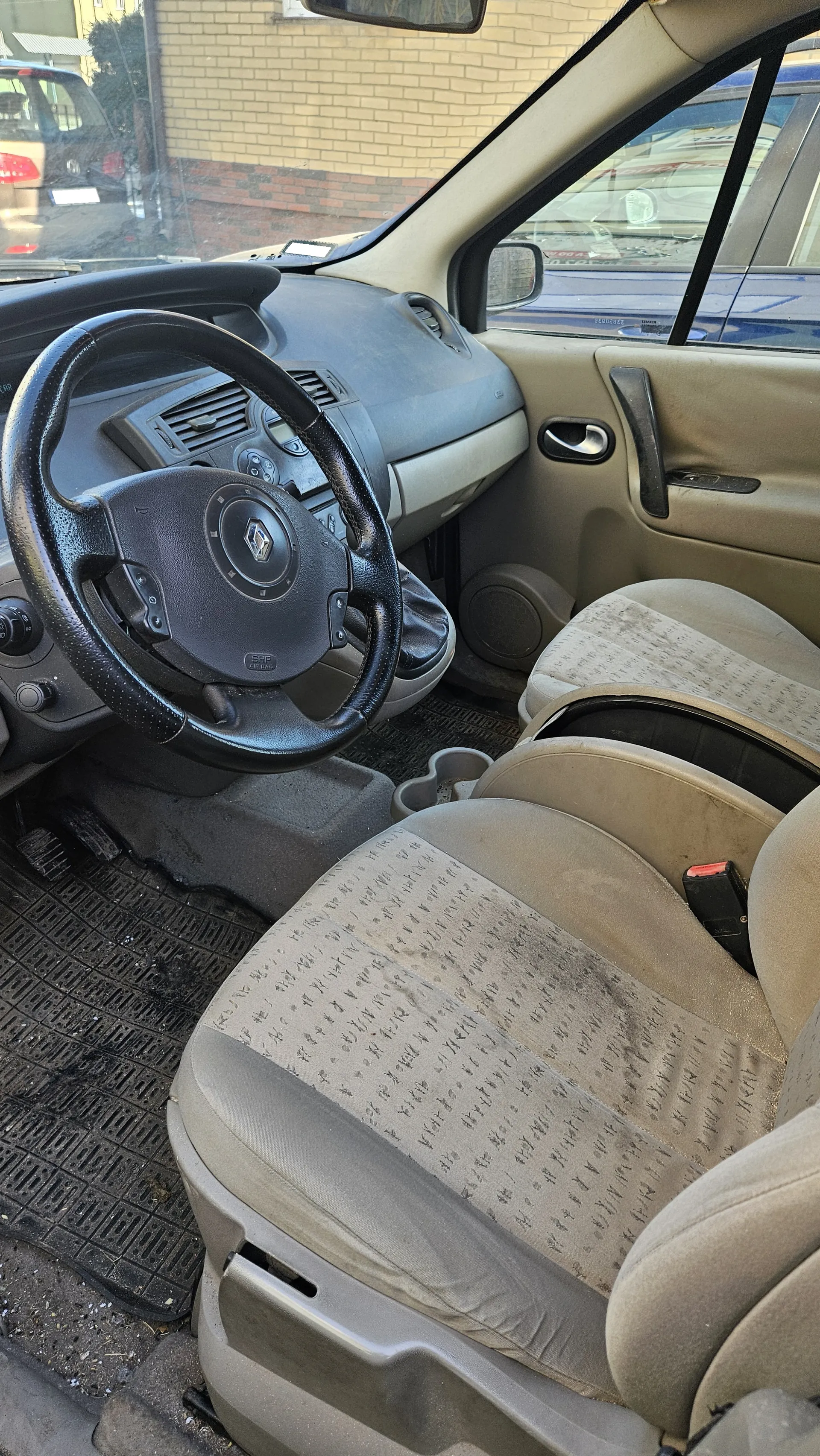 Wnętrze samochodu z kierownicą marki Renault, beżowymi siedzeniami w wzór, zanieczyszczonymi podłogami i dywanikami. Widoczne są deska rozdzielcza, pedały oraz zewnętrzny widok przez przednią szybę na ceglaną ścianę i inne pojazdy.