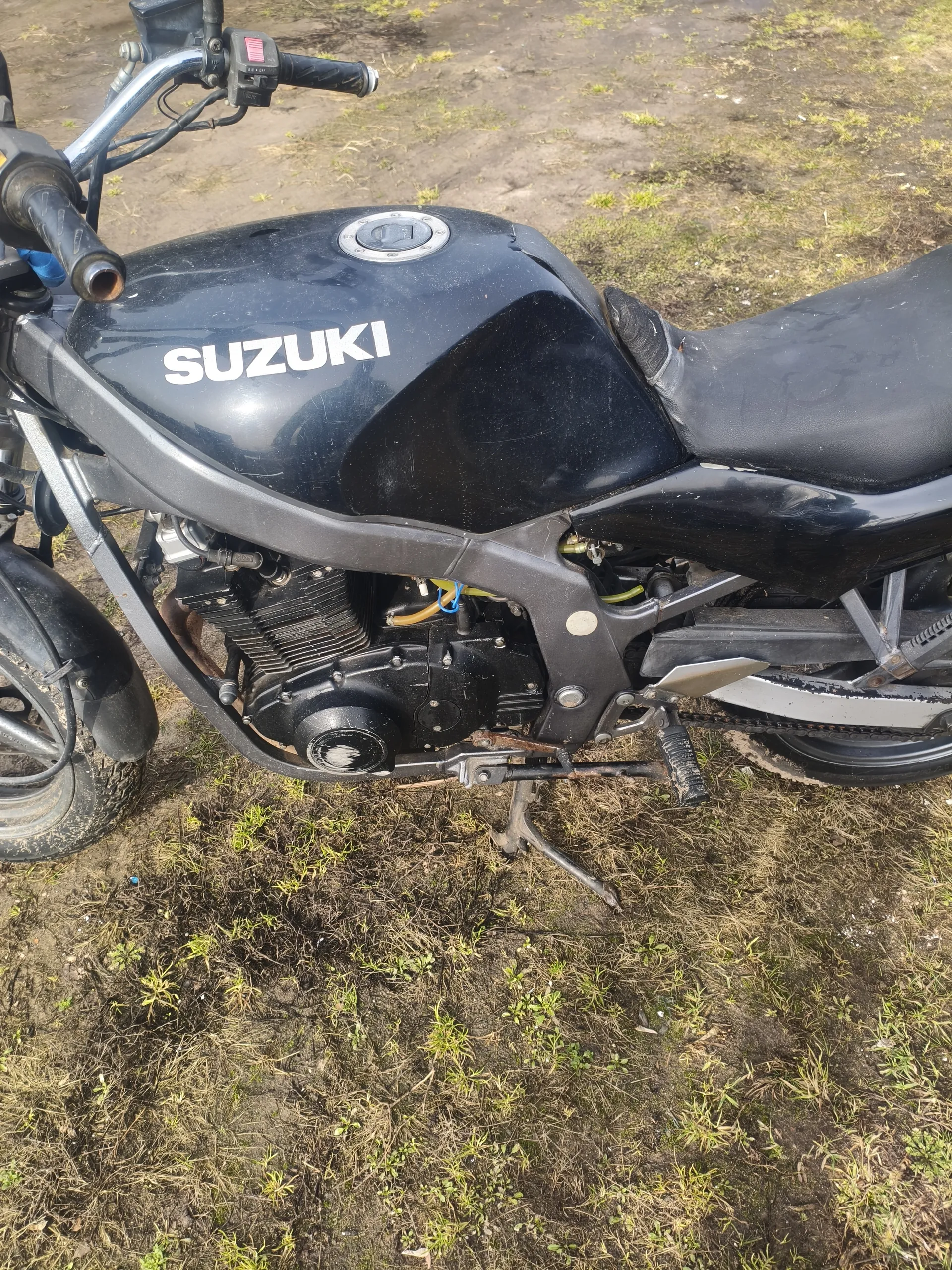 Czarny motocykl marki SUZUKI z widocznym białym napisem na zbiorniku paliwa. Widoczne są elementy takie jak silnik, siedzenie, kierownica z gałką przyspieszenia oraz stojak. Motocykl stoi na trawiastym i błotnistym terenie.