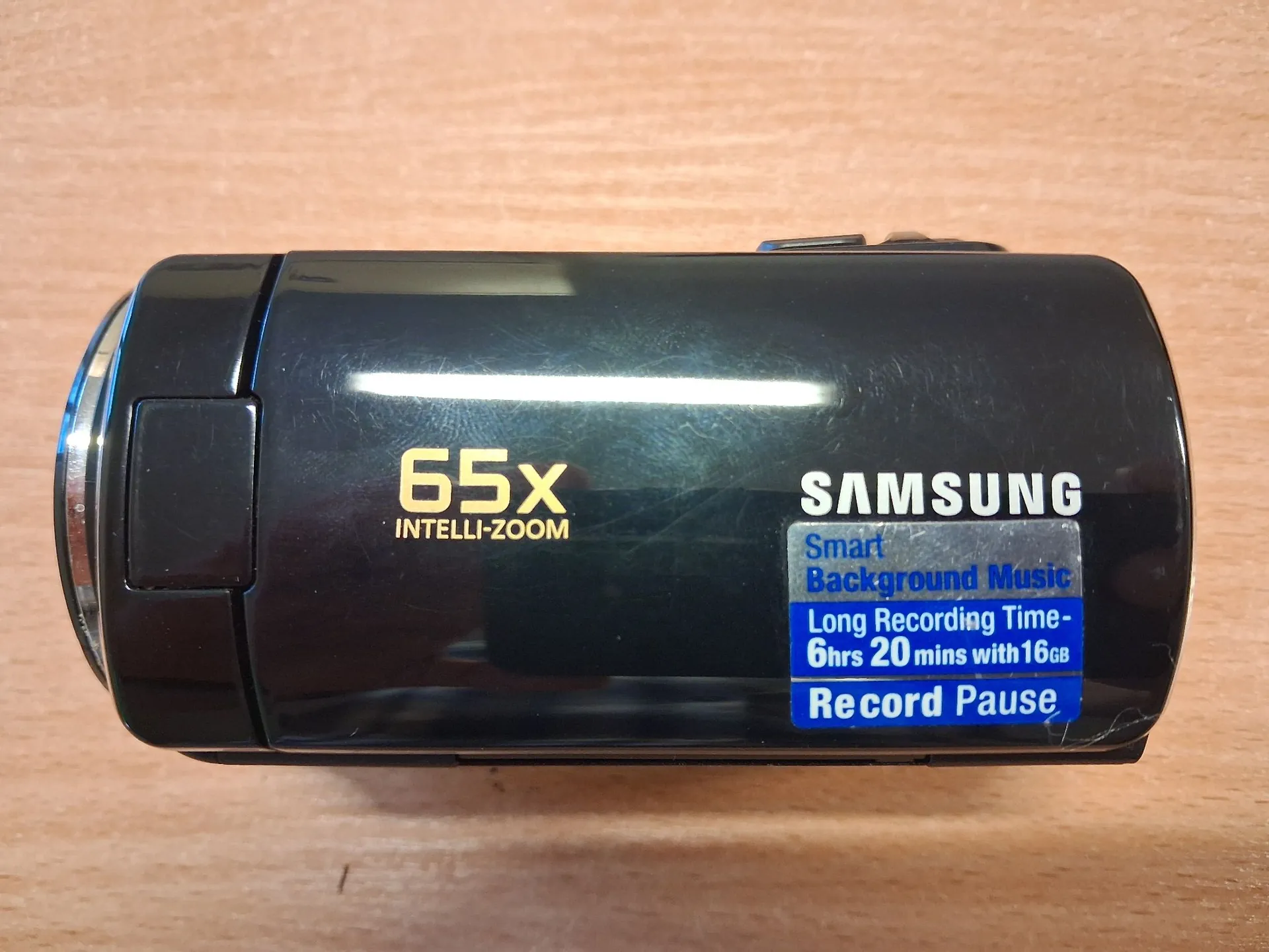 Czarny cyfrowy kamcorder marki Samsung leżący na drewnianej powierzchni. Na obudowie urządzenia widnieją białe napisy: '65X INTELLI-ZOOM' oraz logo 'SAMSUNG'. Obok logo znajduje się naklejka z niebieskim tekstem: 'Smart Background Music', 'Long Recording Time - 6hrs 20 mins with 16GB' oraz 'Record Pause'.