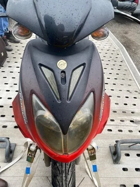 Przód czerwono-czarnego skutera z dużymi reflektorami, pomarańczowymi kierunkowskazami i okrągłym emblematem na czarnej części. Skuter jest przytwierdzony paskami z niebieskimi etykietami do metalowej platformy z otworami. W tle widoczna część nogi osoby w niebieskich spodniach i butach oraz fragment innego pojazdu. Na boku skutera widoczny napis 'Scooter'.