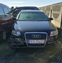 Czarny samochód marki Audi z widocznymi uszkodzeniami przedniej części. Na masce widoczny jest logo Audi, a na tablicy rejestracyjnej napis PKN 0562G. W tle znajdują się inne pojazdy, w tym czerwony samochód po lewej stronie oraz ciemny samochód za Audi. Po prawej stronie obrazu widoczny jest betonowy płot. Tło stanowi otwarta przestrzeń z drobnoziarnistym podłożem.