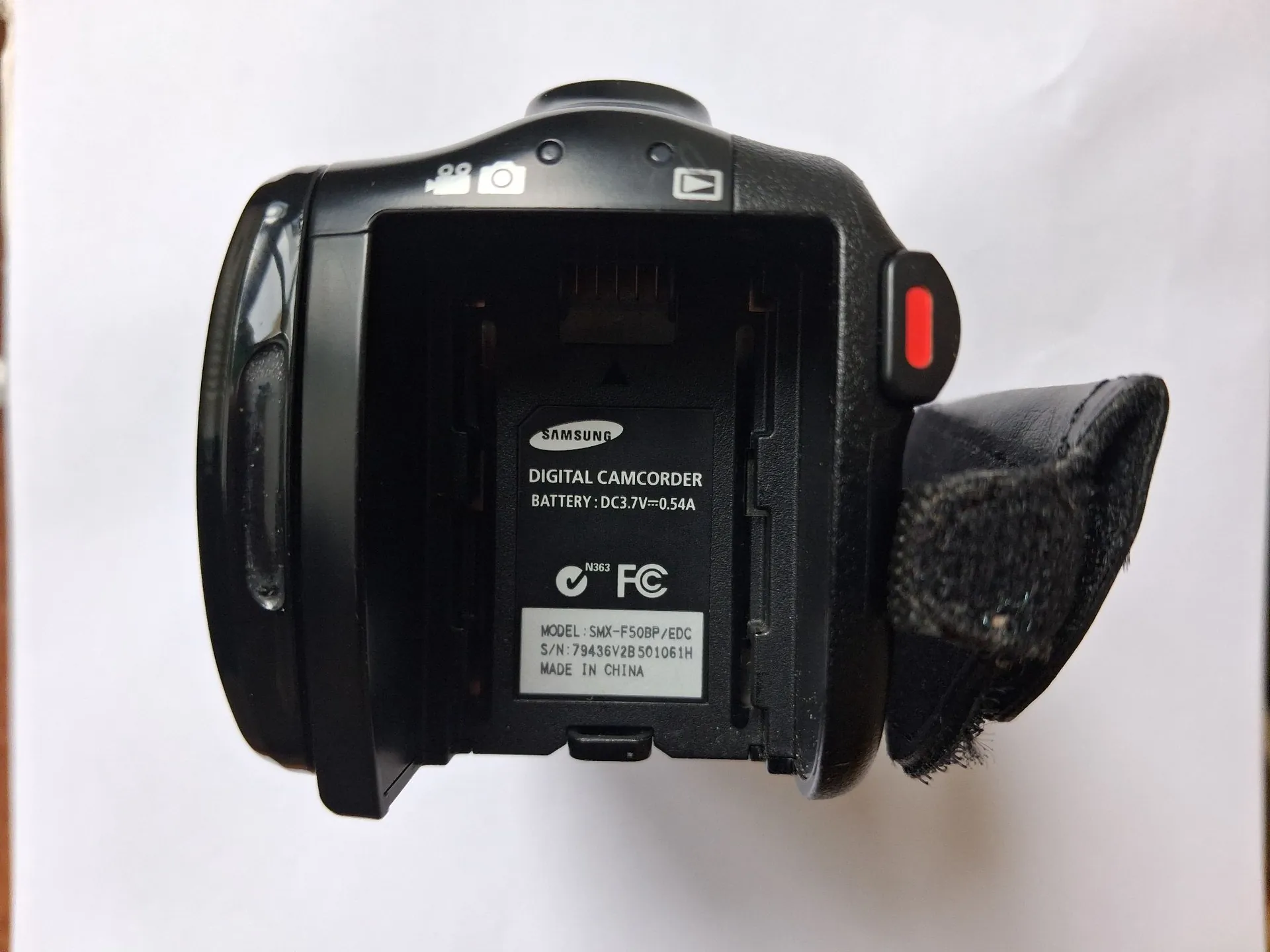 Widok od tyłu czarnej kamery cyfrowej Samsung z otwartą klapką komory baterii. Wewnątrz komory znajduje się biała etykieta z tekstem: 'SAMSUNG DIGITAL CAMCORDER BATTERY: DC3.7V=0.54A', logo FC oraz dane techniczne: 'MODEL: SMX-F50BP/EDC S/N: 79436V2B 501061H MADE IN CHINA'. Po prawej stronie widoczny jest czarny pasek z materiału oraz czerwony przycisk.