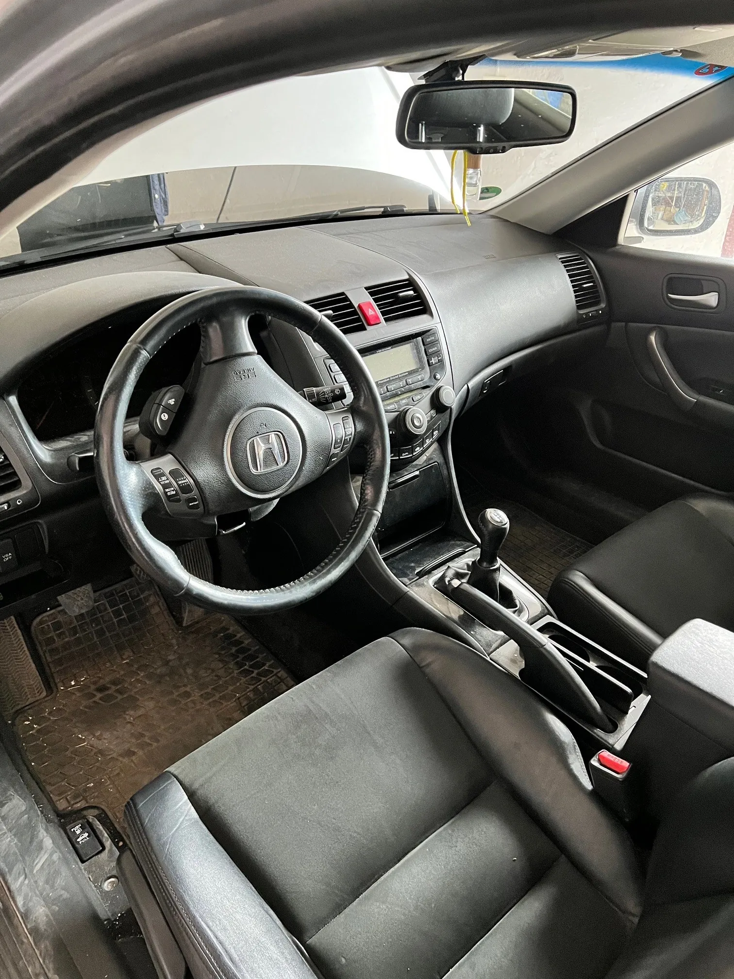 Wnętrze samochodu marki Honda z widocznym kierownicą z logo Hondy, czarną tapicerką, deska rozdzielcza z ekranem, przyciskami i wentylacją, dźwignią zmiany biegów, lusterkiem wstecznym oraz czarnymi dywanikami na podłodze. Widoczne są również czerwone przyciski i elementy sterowania na kierownicy oraz desce rozdzielczej.