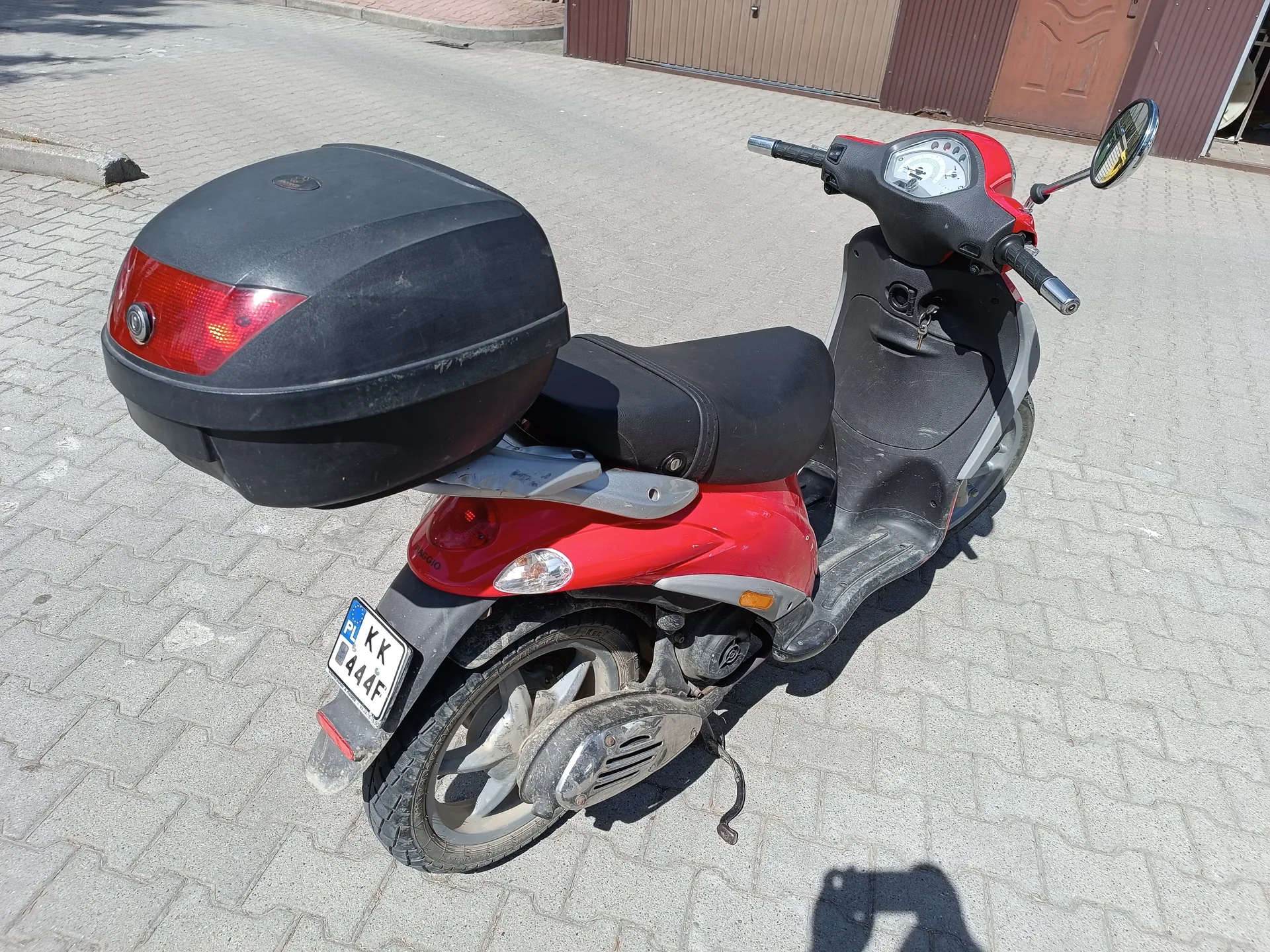 Czerwono-czarny skuter zaparkowany na brukowanej powierzchni. Skuter posiada czarny bagażnik na tylnej części, czerwone światła tylnego oraz tablicę rejestracyjną z napisem PL KK 444F. W tle widoczne są brązowe drzwi garażowe i fragment budynku. Skuter ma czarne siedzenie, kierownicę z lusterkami oraz licznik.
