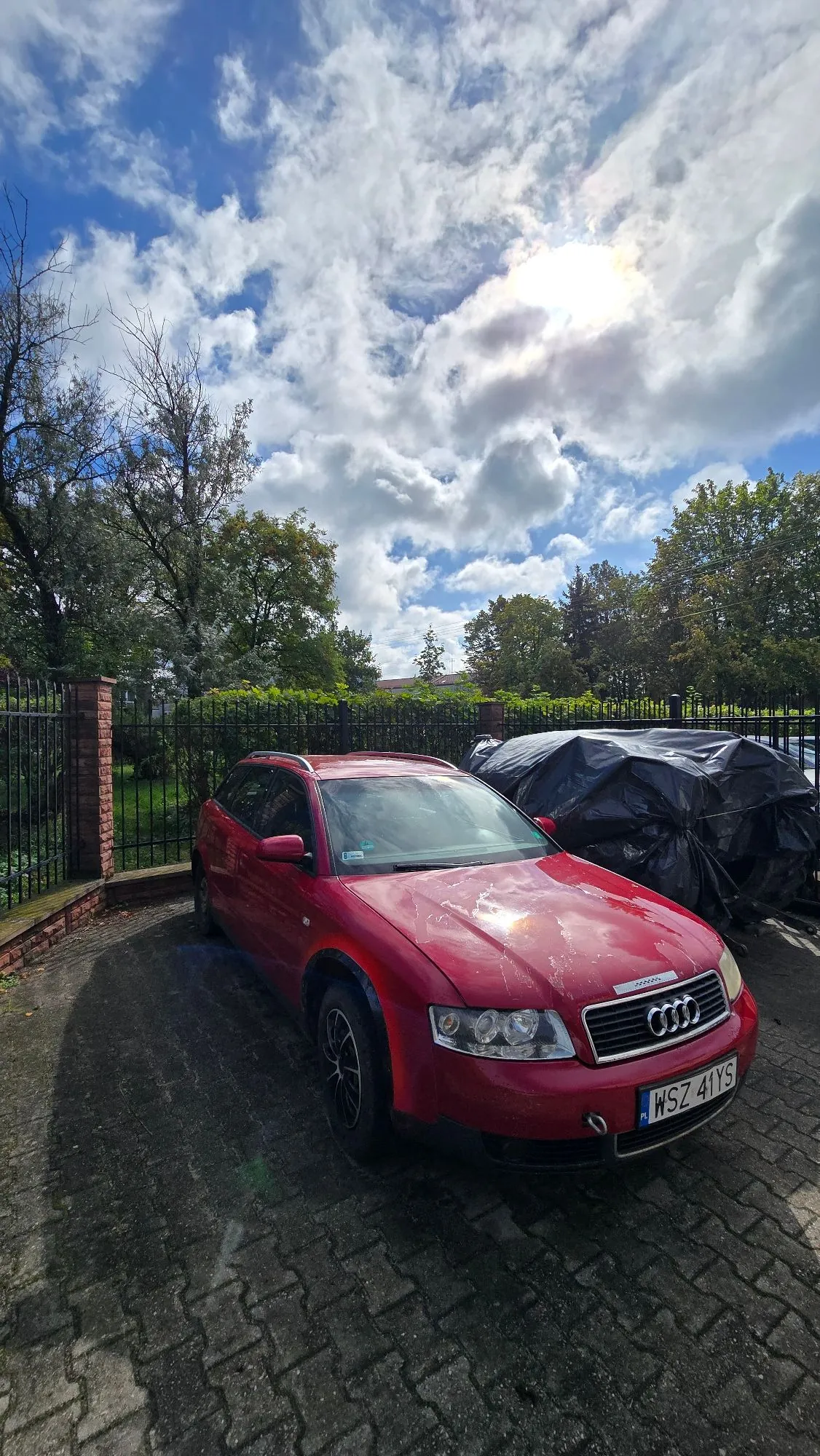 Czerwony samochód marki Audi w nadwoziu kombi zaparkowany na kostce brukowej. Samochód ma widoczne uszkodzenia lakieru na masce. W tle znajduje się czarny płot z ceglanymi słupkami, zielone drzewa oraz inny pojazd przykryty czarną plandeką. Nad wszystkim rozciąga się niebieskie niebo z dużymi, białymi chmurami i widocznym słońcem.