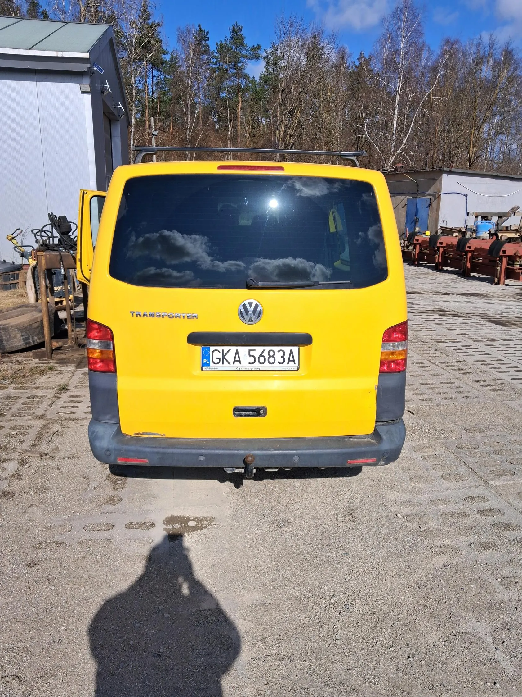 Z tyłu żółty samochód dostawczy Volkswagen Transporter z tablicą rejestracyjną PL GKA 5683A. Po lewej stronie otwarte drzwi, na tle budynki, drzewa i niebo z chmurami. Na asfalcie widoczny cień osoby robiącej zdjęcie. W tle po prawej stronie czerwone konstrukcje metalowe.