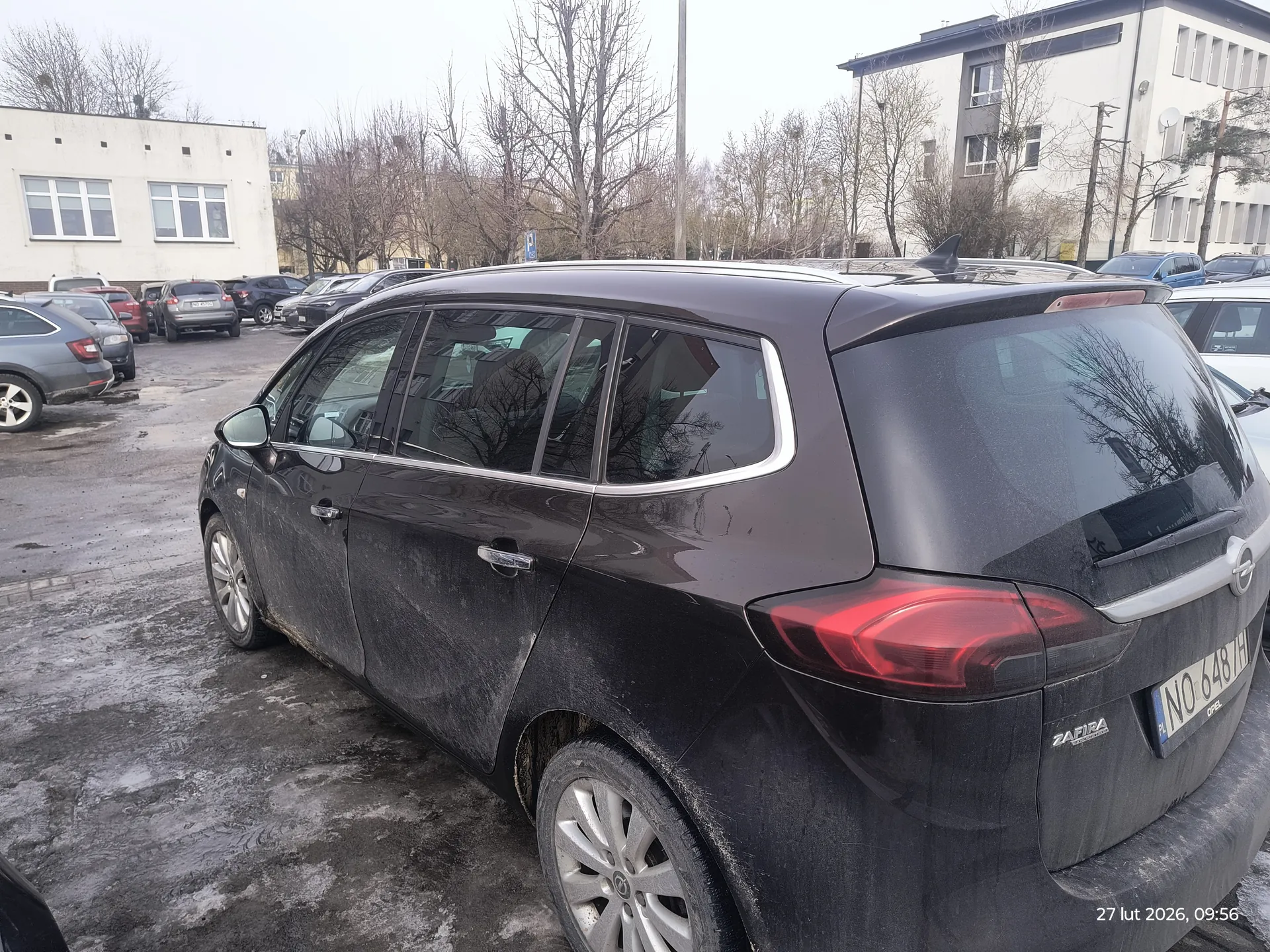 Czarny samochód Opel Zafira zaparkowany na mokrym, częściowo zlodowaciałym parkingu. Pojazd jest zabrudzony, posiada dachowy bagażnik i tablicę rejestracyjną NO 648 TH. W tle widoczne są inne samochody, białe budynki z oknami oraz drzewa bez liści. W prawym dolnym rogu obrazu znajduje się znacznik czasu: 27 lut 2026, 09:56. Na tylnym zderzaku widoczny napis ZAFIRA.
